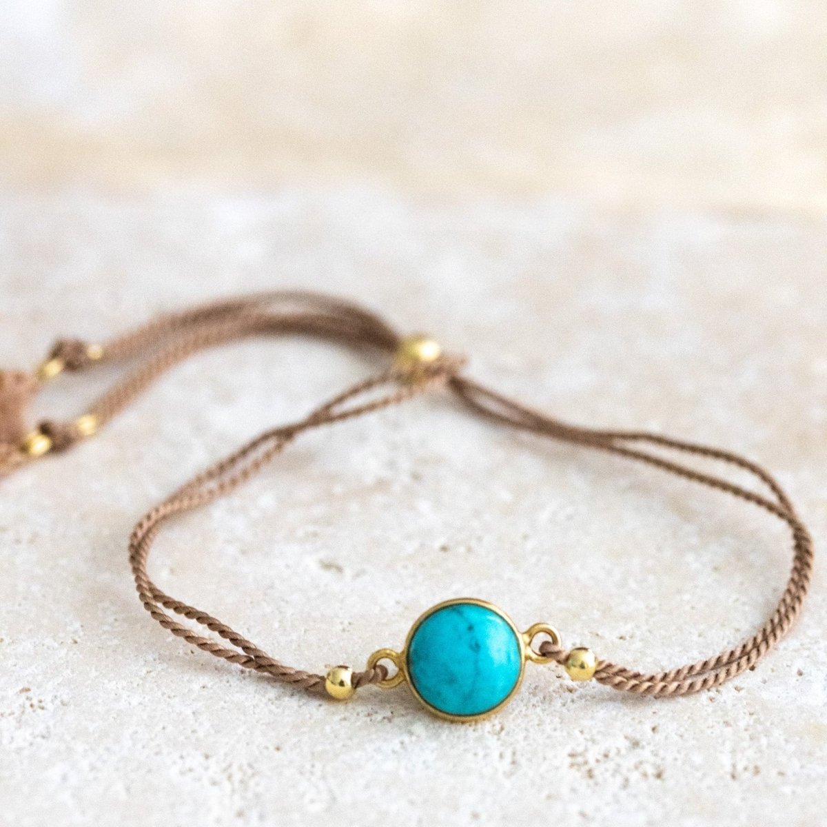 Ein beiges Seidenarmband mit einem runden, gold gefassten Türkis im Zentrum liegt auf einer hellen Natursteinplatte.