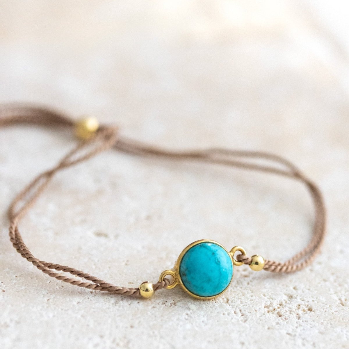 Ein beiges Seidenarmband mit einem runden, gold gefassten Türkis im Zentrum liegt auf einer hellen Natursteinplatte.