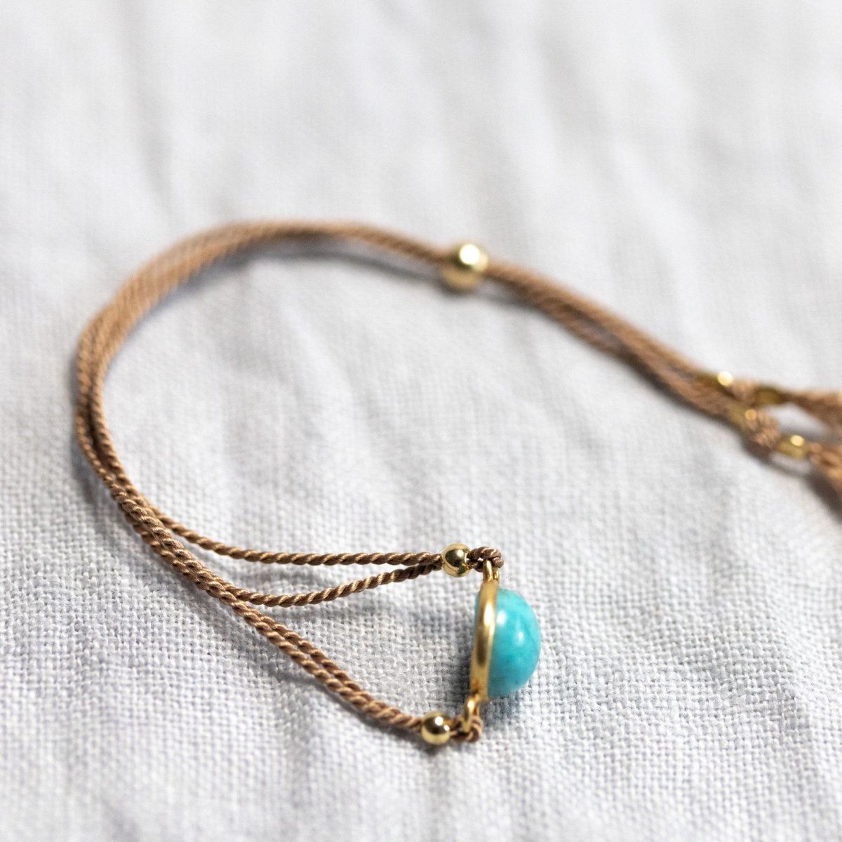 Ein einseitig gewölbter Türkis ist in ein beiges Seidenarmband eingearbeitet. Das Armband liegt auf einem hellen Leinenstoff.