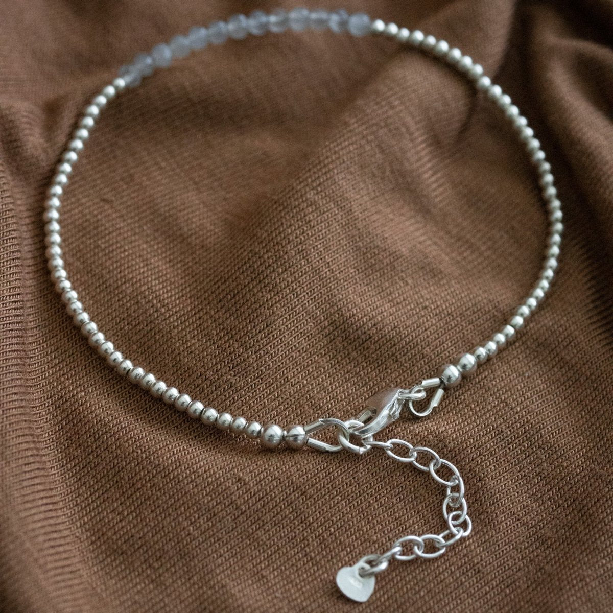 Ein ultra dünnes Silberarmband mit grauen Mondsteinperlen in der Mitte liegt auf braunem Stoff.