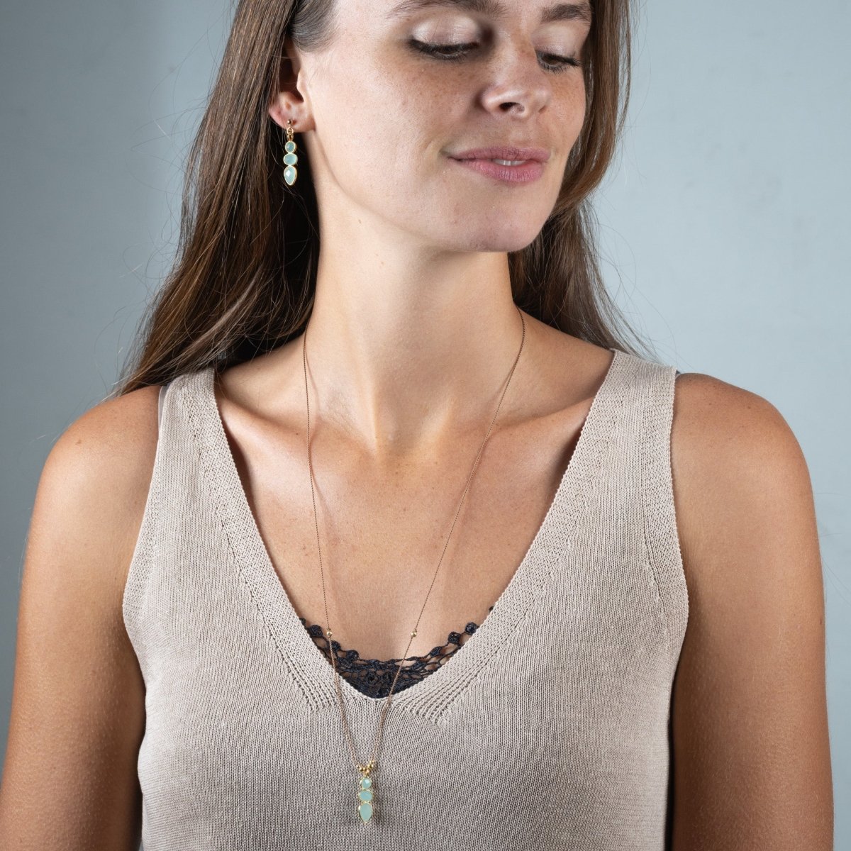 Eine lange Schnurkette mit einem länglichen Anhänger, der aus drei übereinander in gold gefassten Aqua Chalcedonen besteht, hängt am Hals des Models.