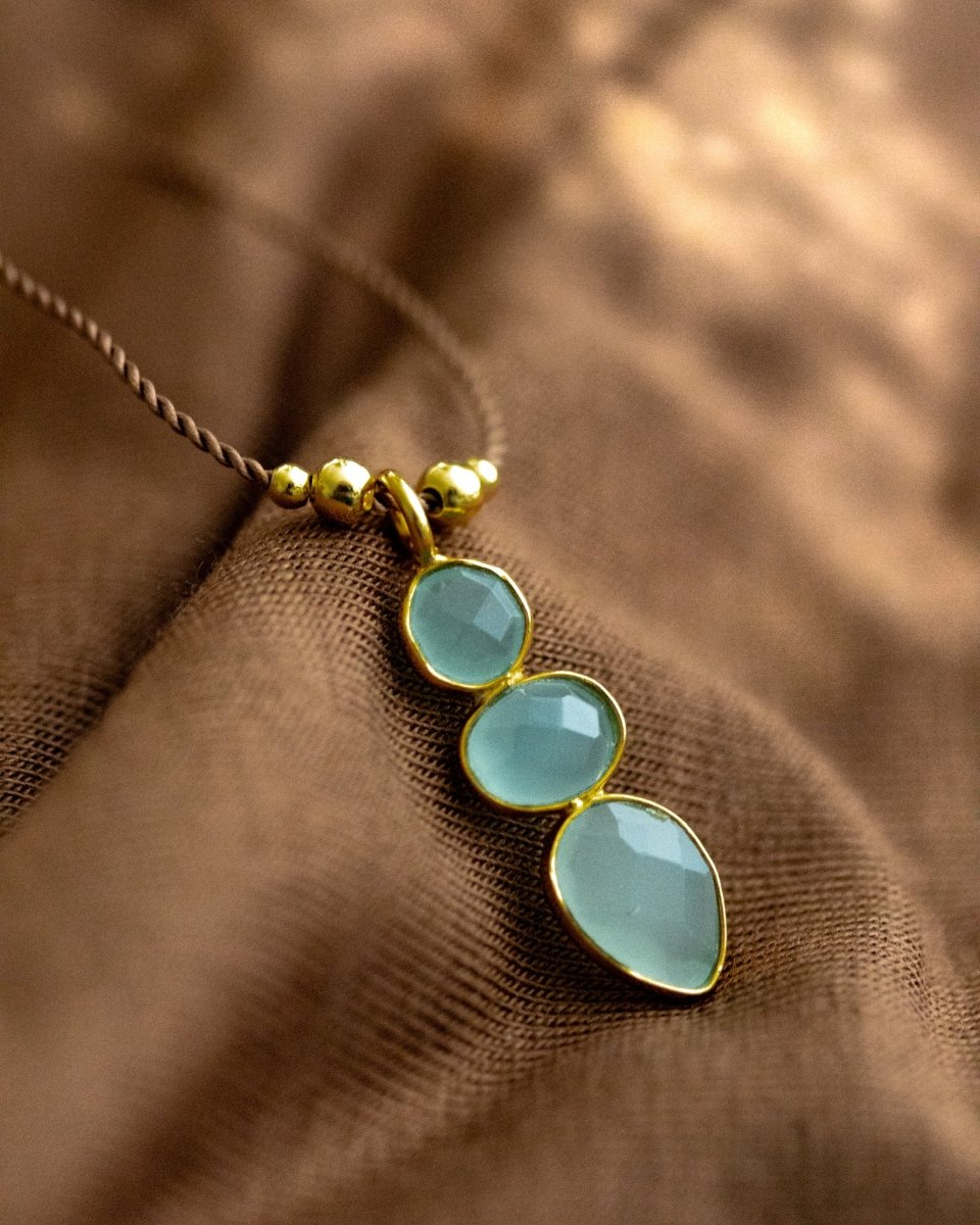 Eine lange Schnurkette mit einem länglichen Anhänger, der aus drei übereinander in gold gefassten Aqua Chalcedonen besteht, liegt auf braunem Stoff.