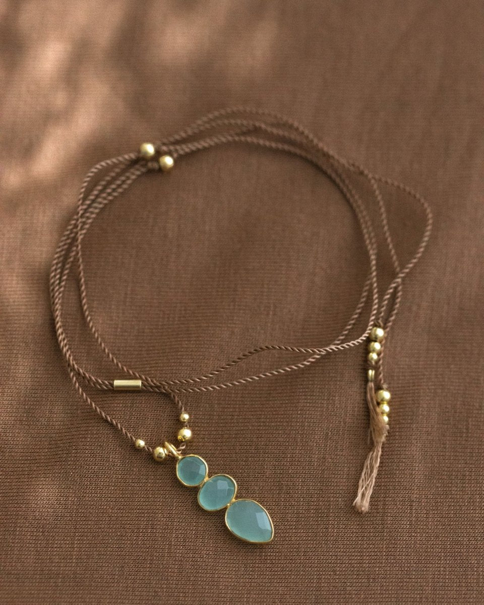 Eine lange Schnurkette mit einem länglichen Anhänger, der aus drei übereinander in gold gefassten Aqua Chalcedonen besteht, liegt auf braunem Stoff.