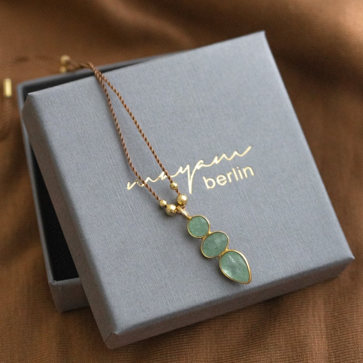 Eine lange, dünne Schnurkette mit einem länglichen Anhänger, der aus drei übereinander in gold gefassten Prehnit Edelsteinen besteht, liegt auf einem grauen Schmucketui.