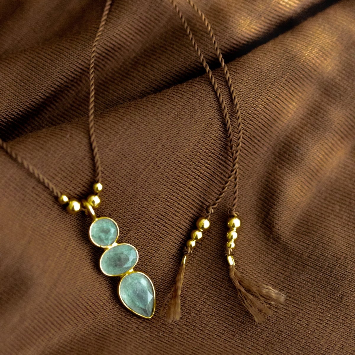 Eine lange, dünne Schnurkette mit einem länglichen Anhänger, der aus drei übereinander in gold gefassten Prehnit Edelsteinen besteht, liegt auf braunem Stoff.