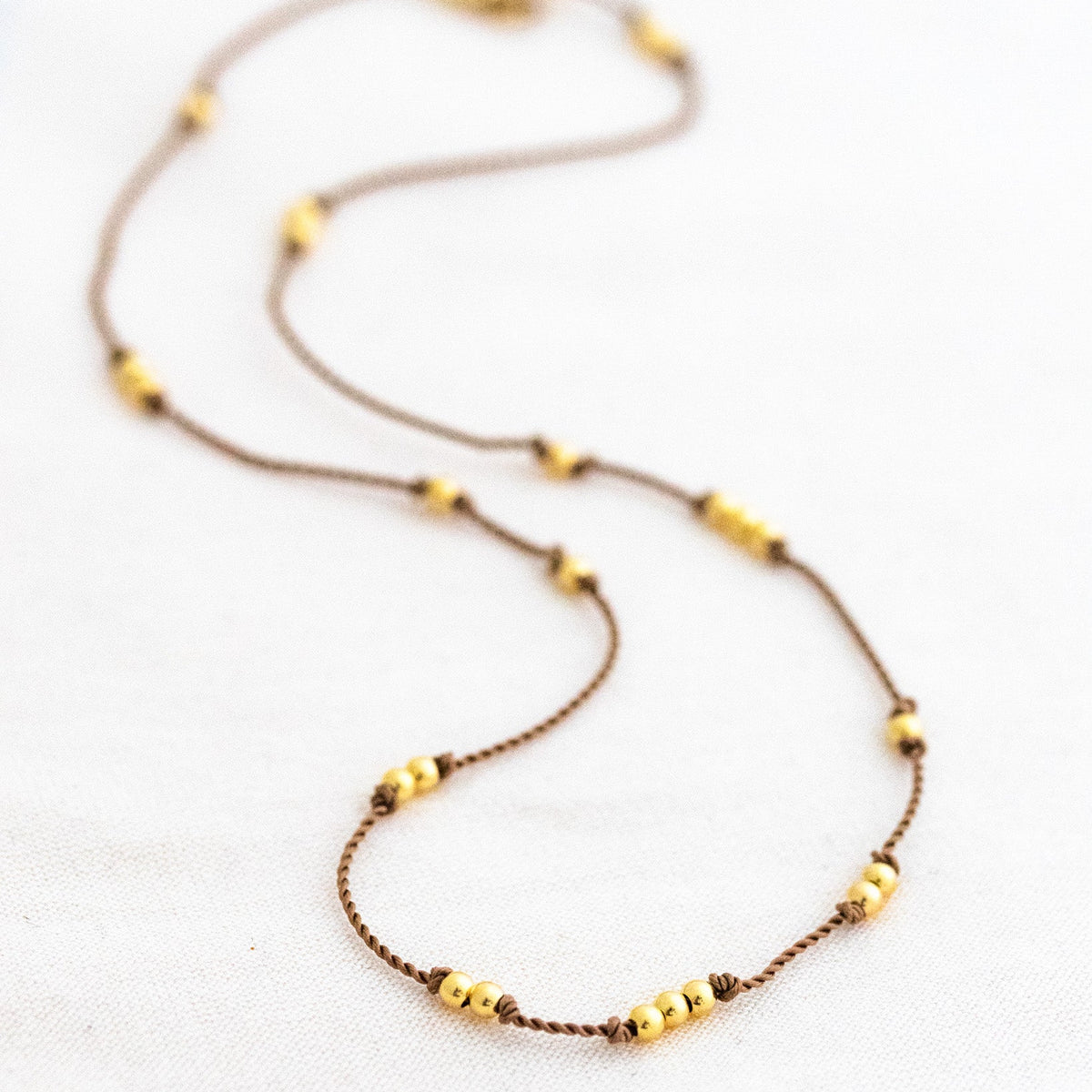 Dünne, beige Kordelkette mit unregelmäßig verteilten Goldperlen ausgelegt auf weißem Untergrund.