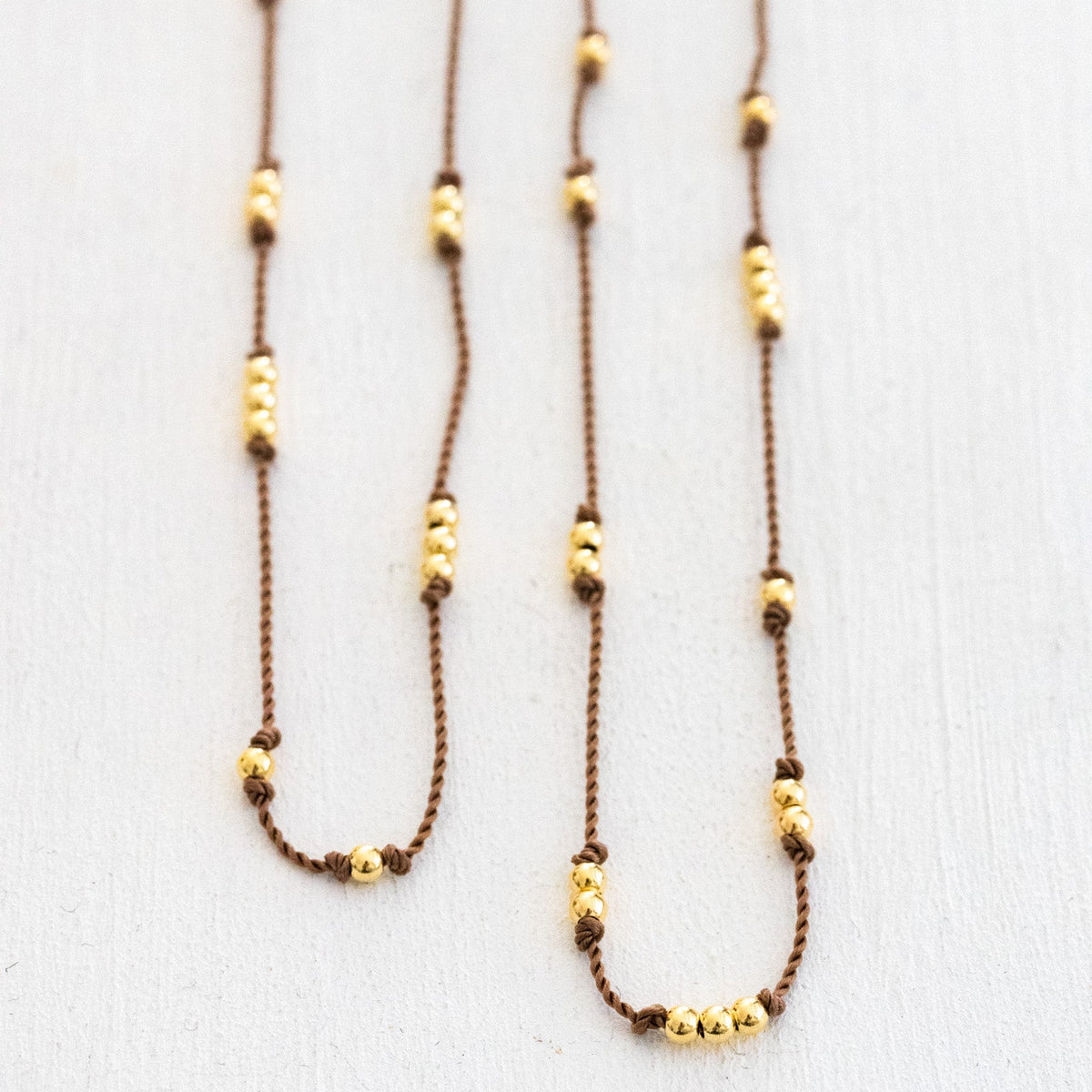 Zwei dünne, beige Kordelketten mit unregelmäßig verteilten Goldperlen auf weißer Fläche ausgelegt.