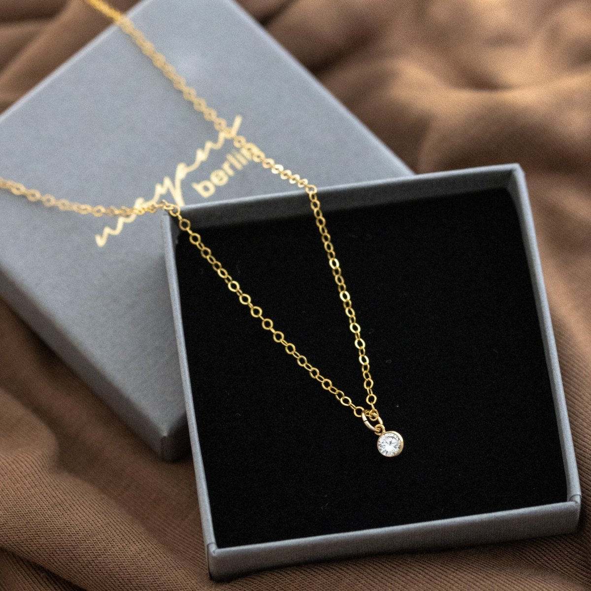 Eine feine Goldkette mit einem winzigen, runden Zirkonia-Anhänger liegt auf einem geöffneten Schmucketui mit der Beschriftung mayam berlin.