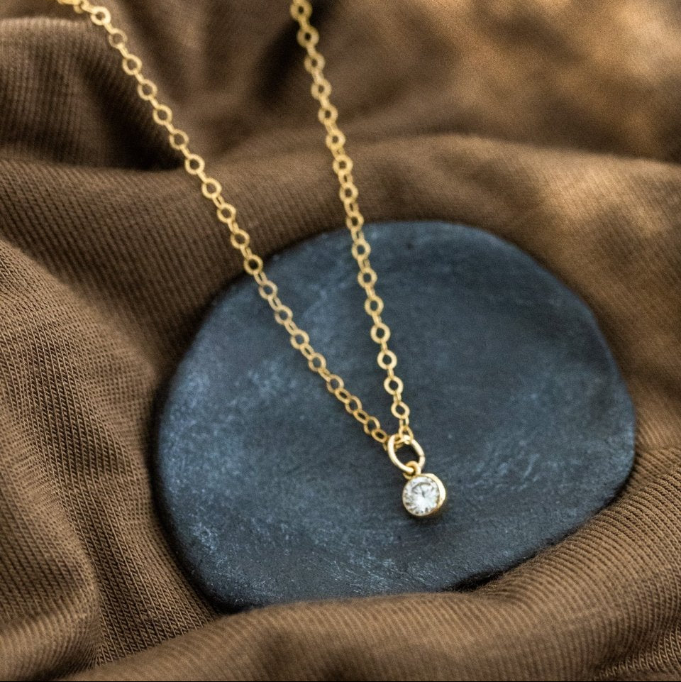 Eine feine Goldkette mit einem winzigen, runden Zirkonia-Anhänger liegt auf einem schwarzen Schälchen auf braunem Stoff.