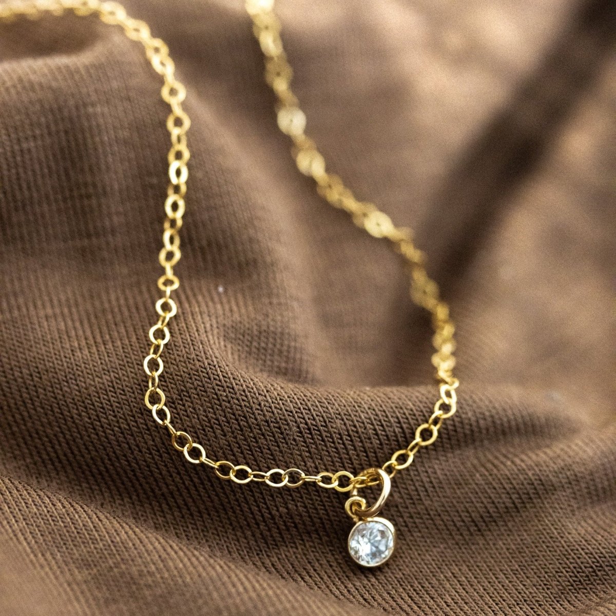 Eine feine Goldkette mit einem winzigen, runden Zirkonia-Anhänger liegt auf gewelltem, braunen Stoff.