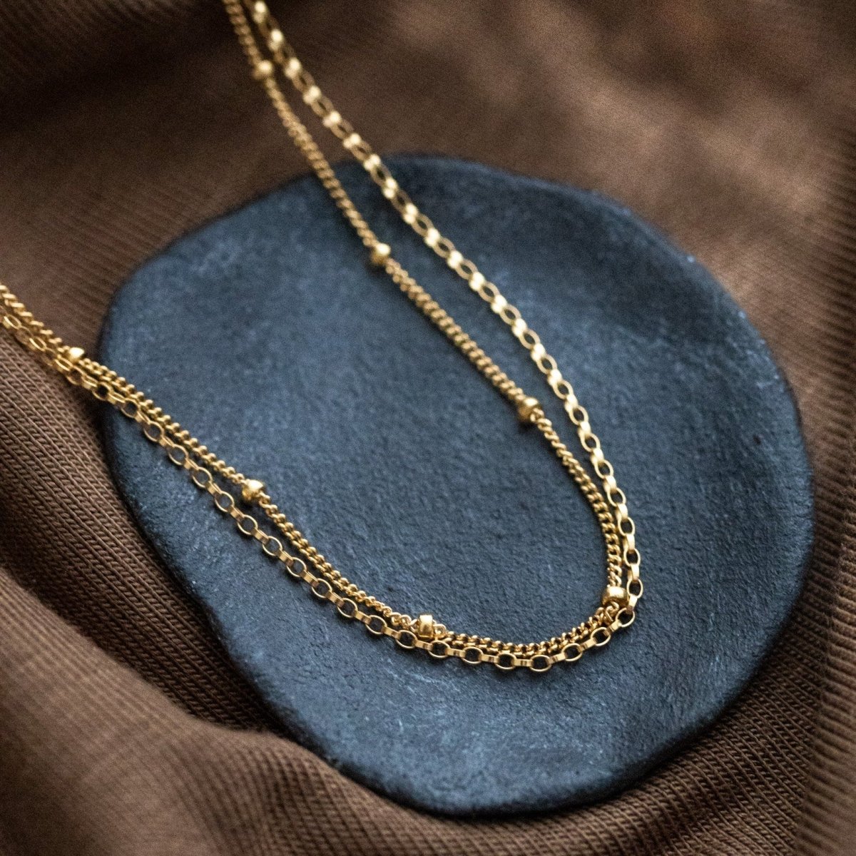 Eine goldene Halskette, bestehend aus einer Erbskette und einer Satellitenkette, liegt auf einem grauen Steinschälchen, das auf braunem Stoff steht.