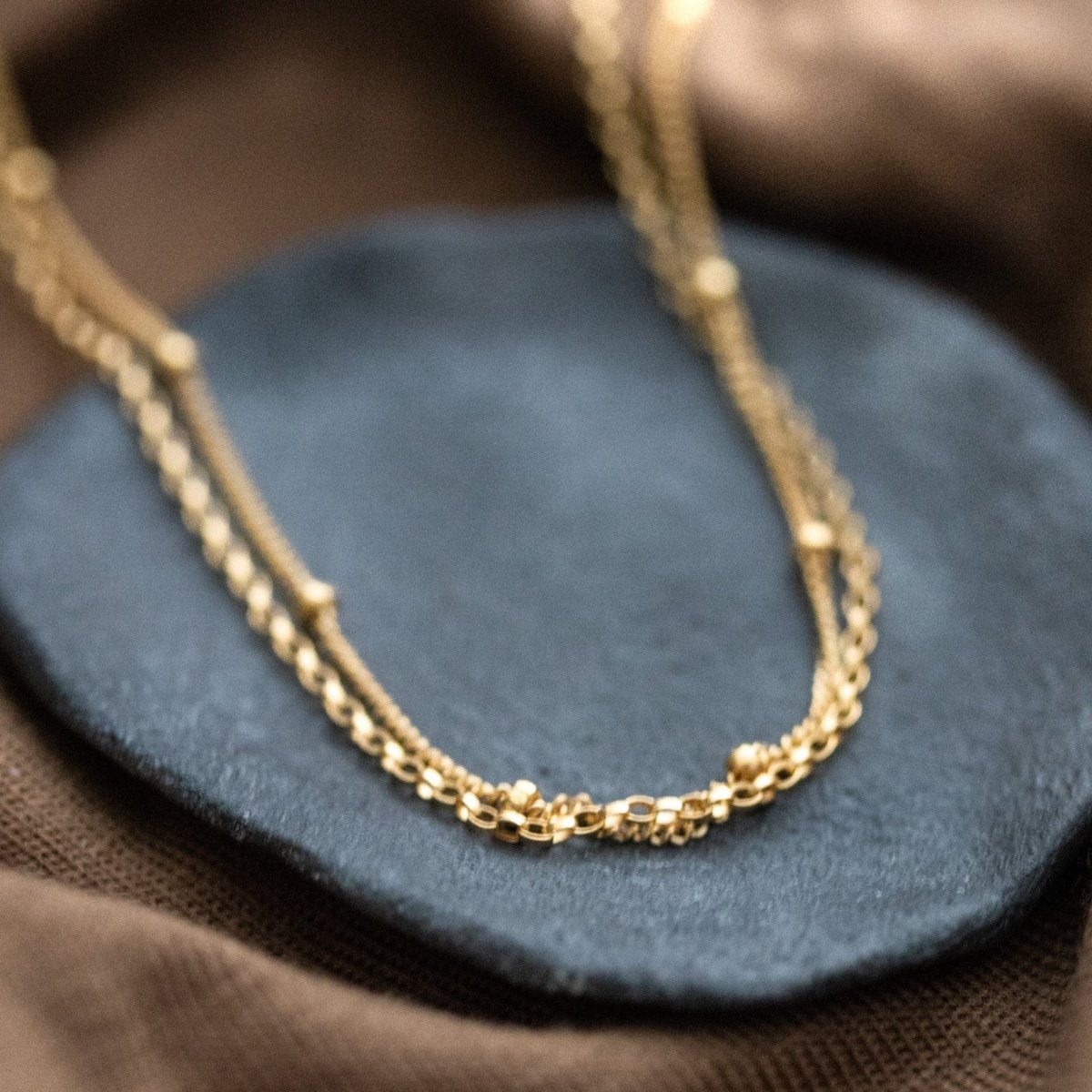 Eine goldene Halskette, bestehend aus einer Erbskette und einer Satellitenkette, liegt auf einem grauen Steinschälchen, das auf braunem Stoff steht.