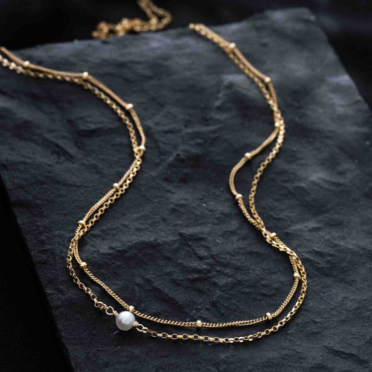 Eine zweireihige Goldkette mit einer kleinen Süßwasserperle auf schwarzem Hintergrund.
