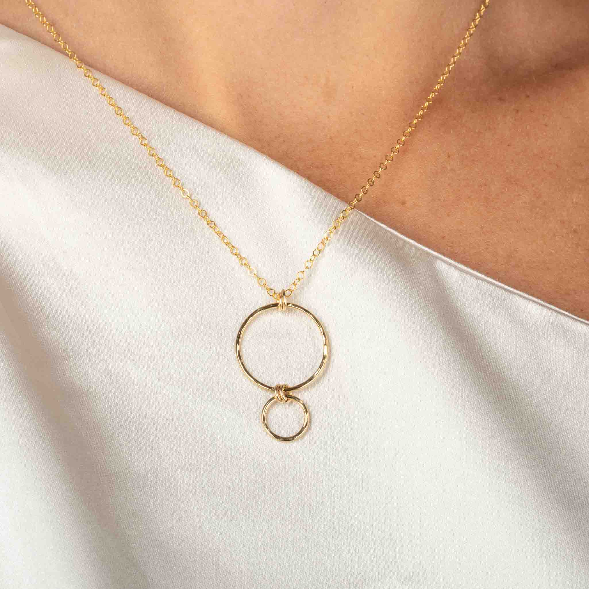 Eine feine Goldkette mit einem Anhänger, bestehend aus einem größeren Ring, an dem ein kleinerer Ring hängt, hängt vom Hals des Models.
