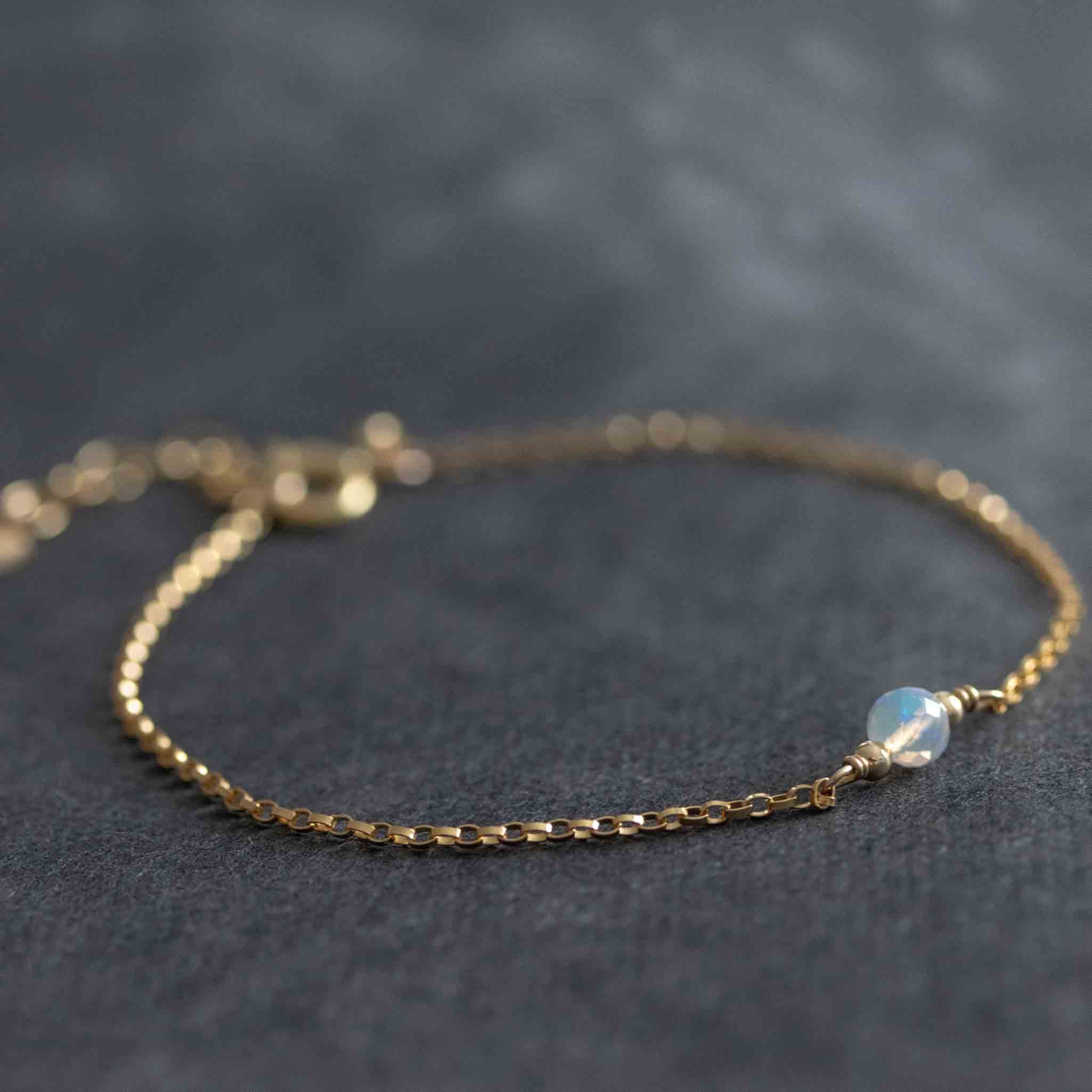 Ein dünnes, goldenes Armband mit einem kleinen Opal Edelstein als Mittelstück liegt auf einem dunklen Hintergrund. 