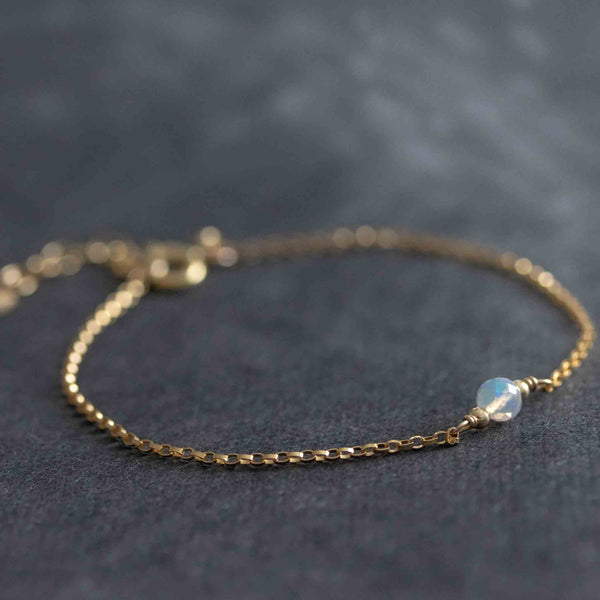 Ein dünnes, goldenes Armband mit einem kleinen Opal Edelstein als Mittelstück liegt auf einem dunklen Hintergrund. 