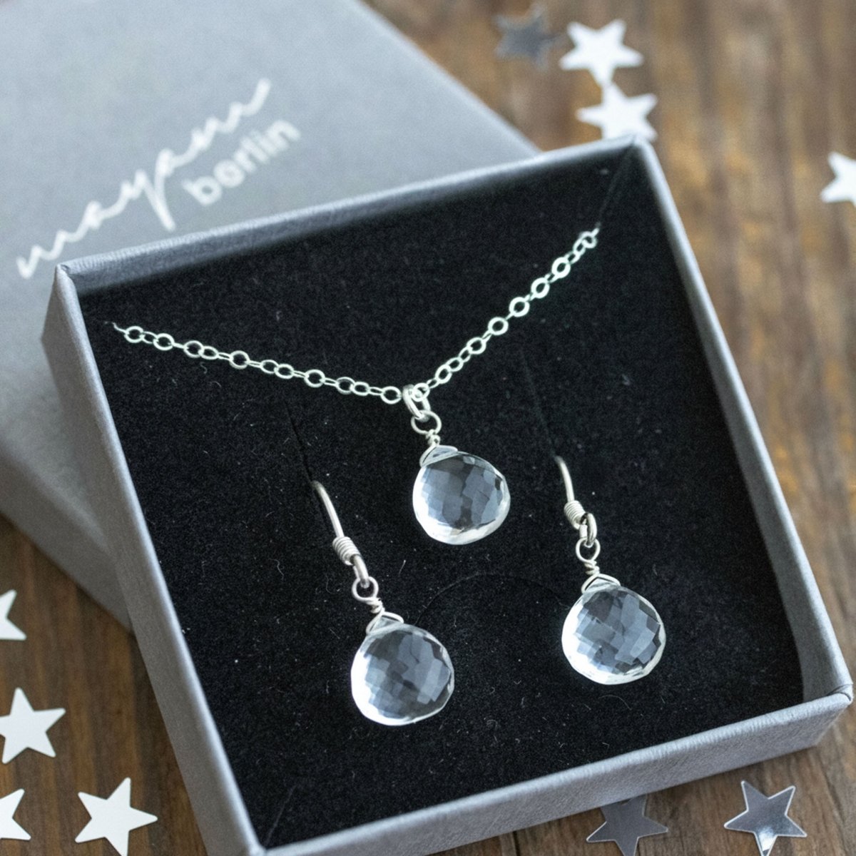 In ein offenes Schmucketui ist eine Silberkette mit Bergristall-Anhänger und ein passendes Paar Ohrring mit dem gleichen Stein eingehängt. Auf dem Holzuntergrund liegt silber-metallisches Sternchenkonfetti verstreut.