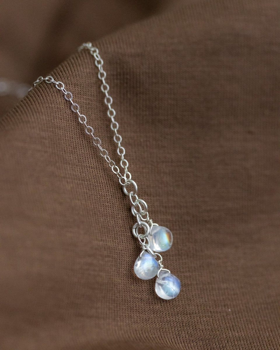 Eine dünne Silberkette mit drei untereinander angebrachten Mondstein-Tropfen lehnt an braunem Stoff.