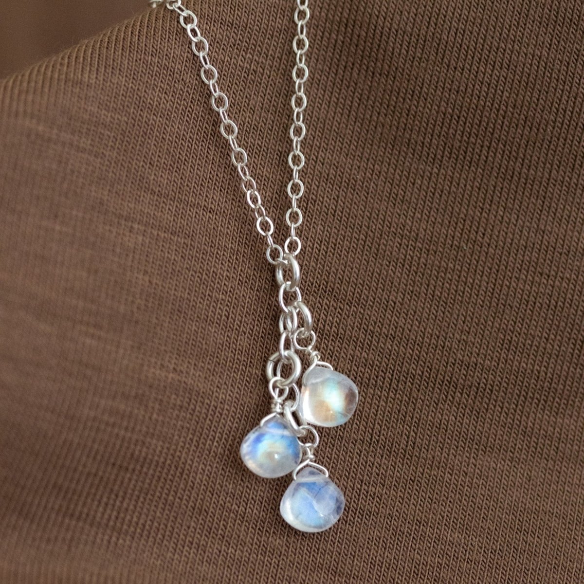 Eine feine Silberkette mit drei untereinander angebrachten Mondstein-Anhängern lehnt an einem braunen Hintergrund.