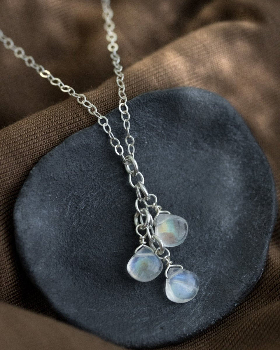 Eine dünne Silberkette mit drei untereinander angebrachten Mondstein-Tropen liegt auf einem grauen Steinschälchen.