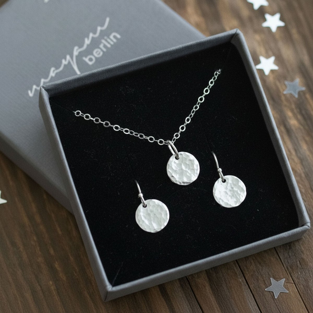 In einem offenen Schmucketui ist ein Set eingehängt, bestehend aus einer Kette und Ohrringen mit jeweils einem runden, gehämmerten Plättchen-Anhänger. Auf dem Holzuntergrund liegt ein wenig Sternchenkonfetti verteilt.