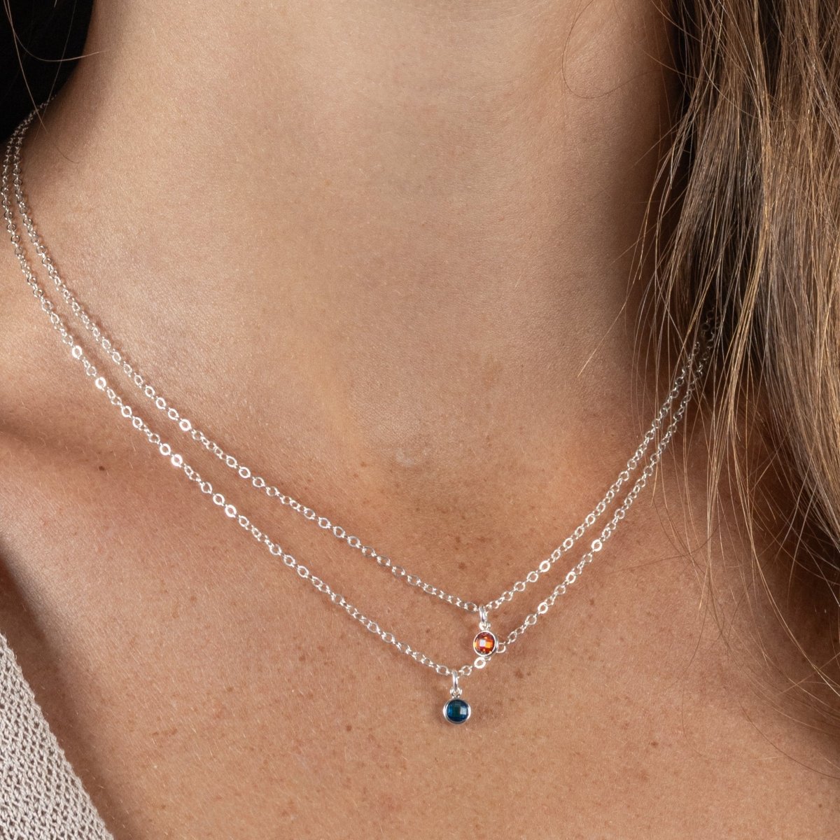 Am Hals des Models hängen zwei filigrane Silberketten mit je einem kleinen, rund gefassten Steinanhänger, einmal in rot und einmal in blau.