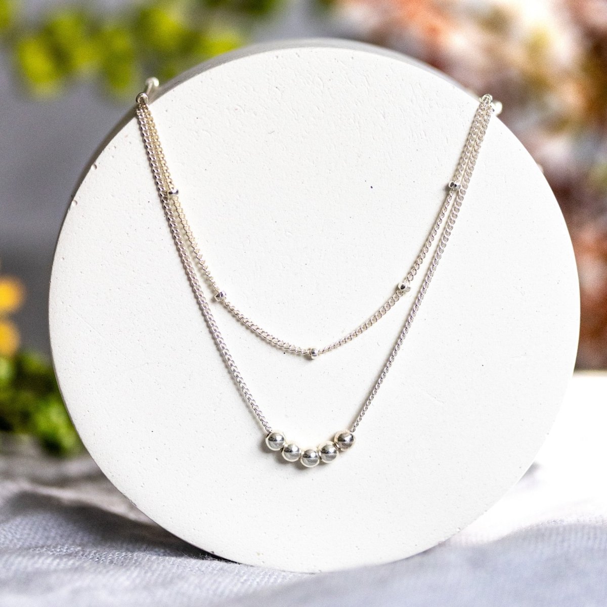 An einer weißen, runden Platte hängen zwei feine Silberketten. Die kürzere hat winzige silberne Akzente, auf die längere sind fünf kleine Silbperlen aufgezogen. Verschwommener, floraler Hintergrund.