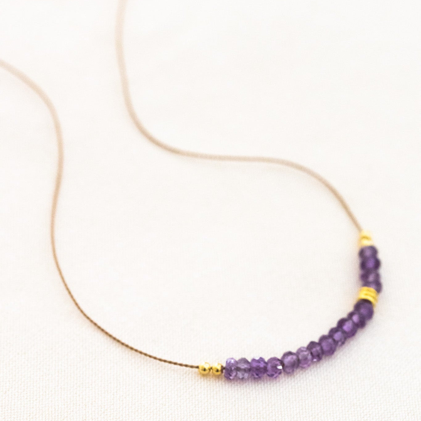 Auf weißen Hintergrund liegt eine kurze Halskette. Sie besteht aus einer dünnen Seidenschnur mit einer Reihe kleiner, violetter Amethyst-Rondellen und goldenen Zierperlen in der Mitte.