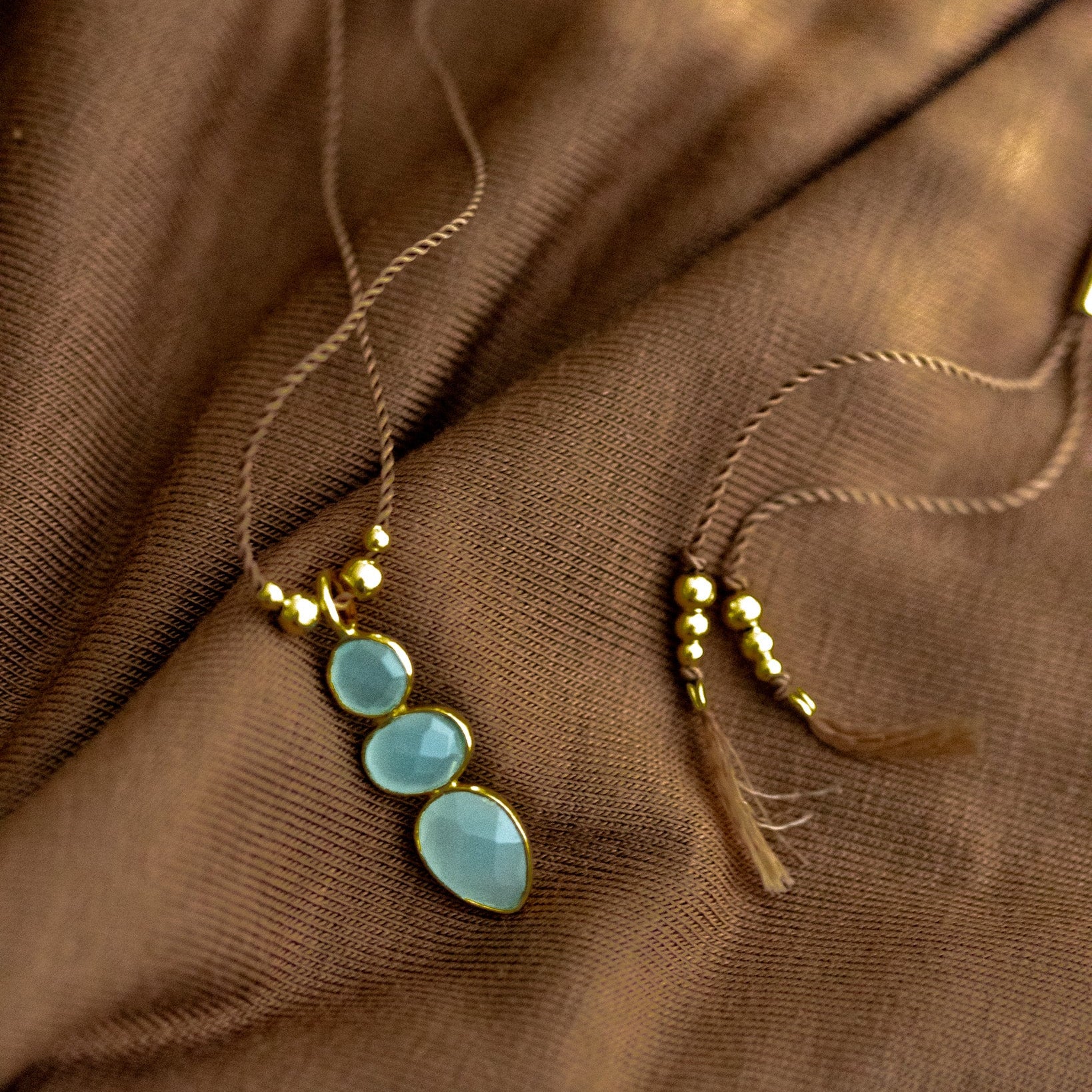 Eine lange Schnurkette mit einem länglichen Anhänger, der aus drei übereinander in gold gefassten Aqua Chalcedonen besteht, liegt auf braunem Stoff.
