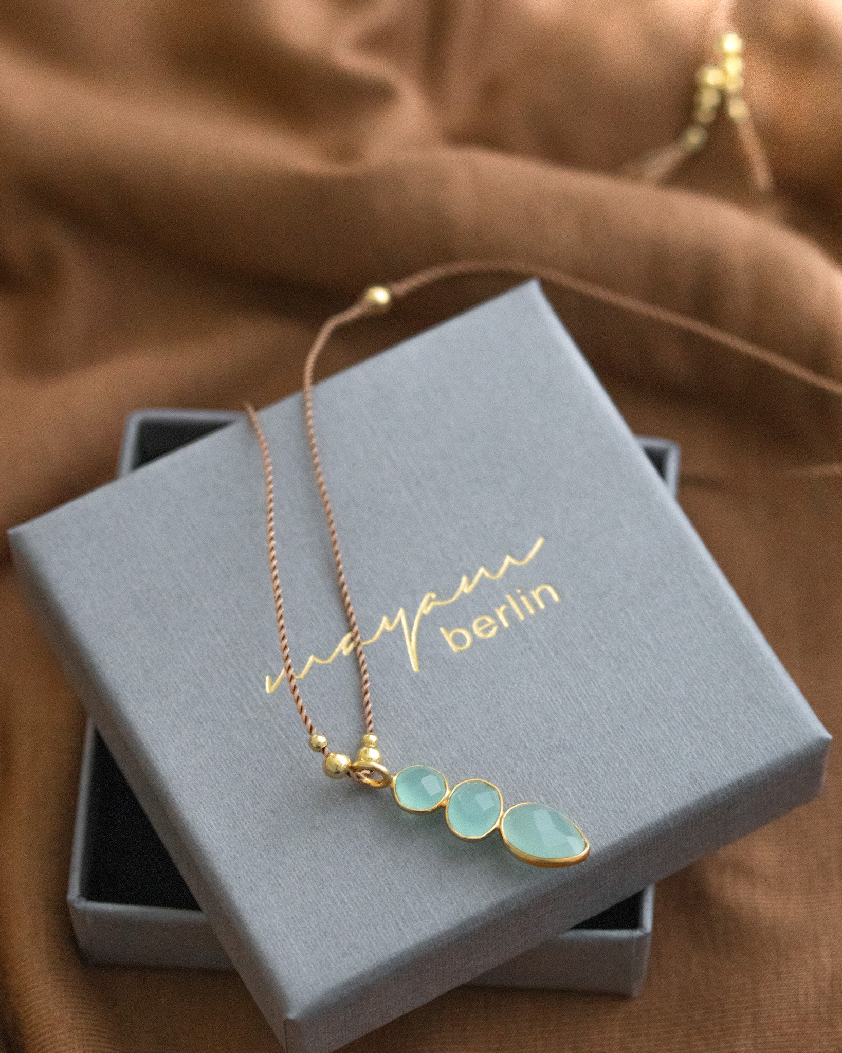 Eine lange Schnurkette mit einem länglichen Anhänger, der aus drei übereinander in gold gefassten Aqua Chalcedonen besteht, liegt auf einem grauen Schmucketui.