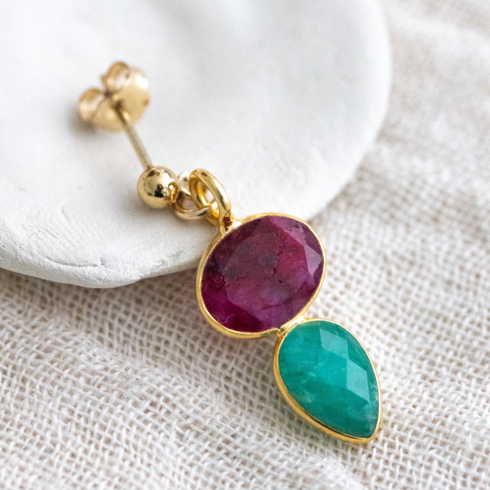 Ein Ohrstecker, der aus einem gold gerahmten Rubin und einem darunter angebrachten, ebenfalls gold gefassten, Smaragd besteht, lehnt an einem weißen Schälchen.