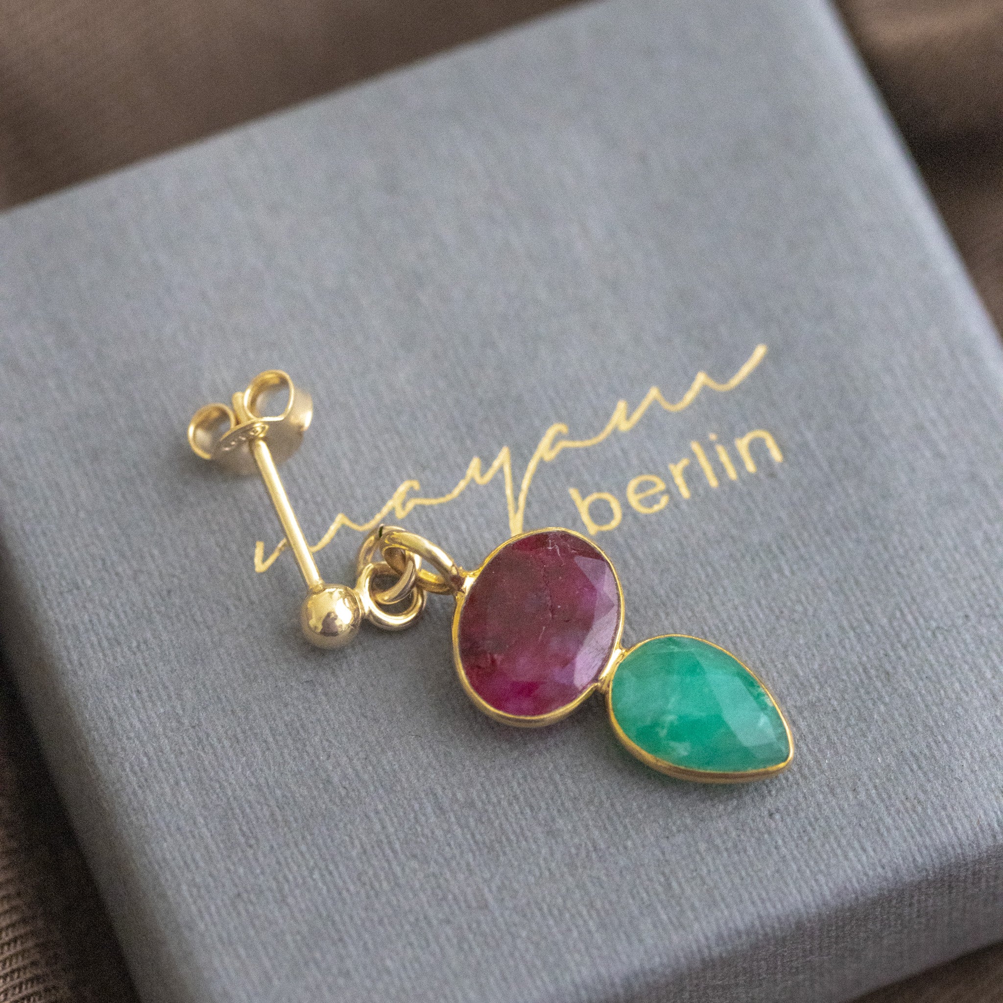 Ein Ohrstecker, der aus einem gold gerahmten Rubin und einem darunter angebrachten, ebenfalls gold gefassten, Smaragd besteht, liegt auf einem grauen Schmucketui.