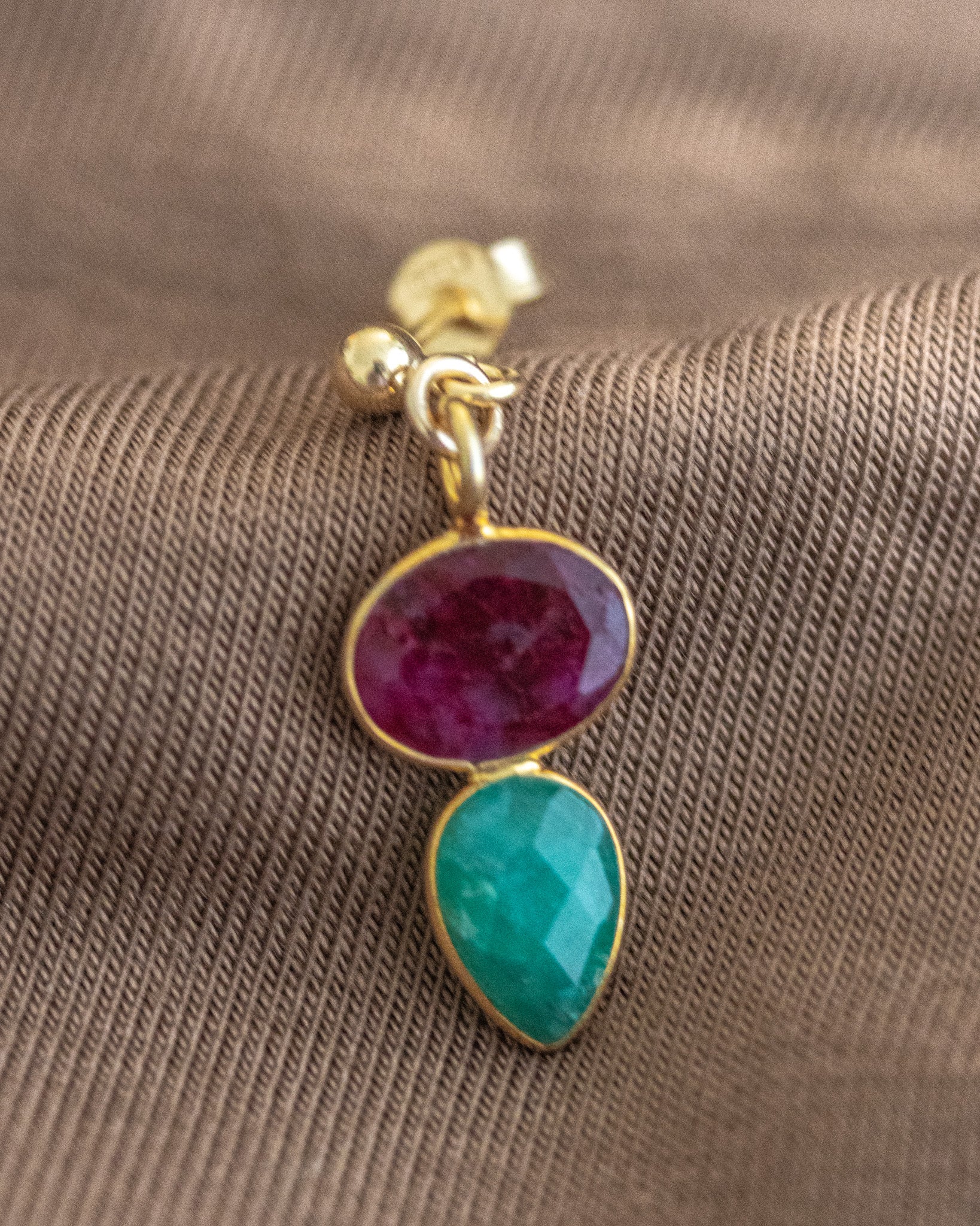 Ein Ohrstecker, der aus einem gold gerahmten Rubin und einem darunter angebrachten, ebenfalls gold gefassten, Smaragd besteht, lehnt an braunem Stoff.