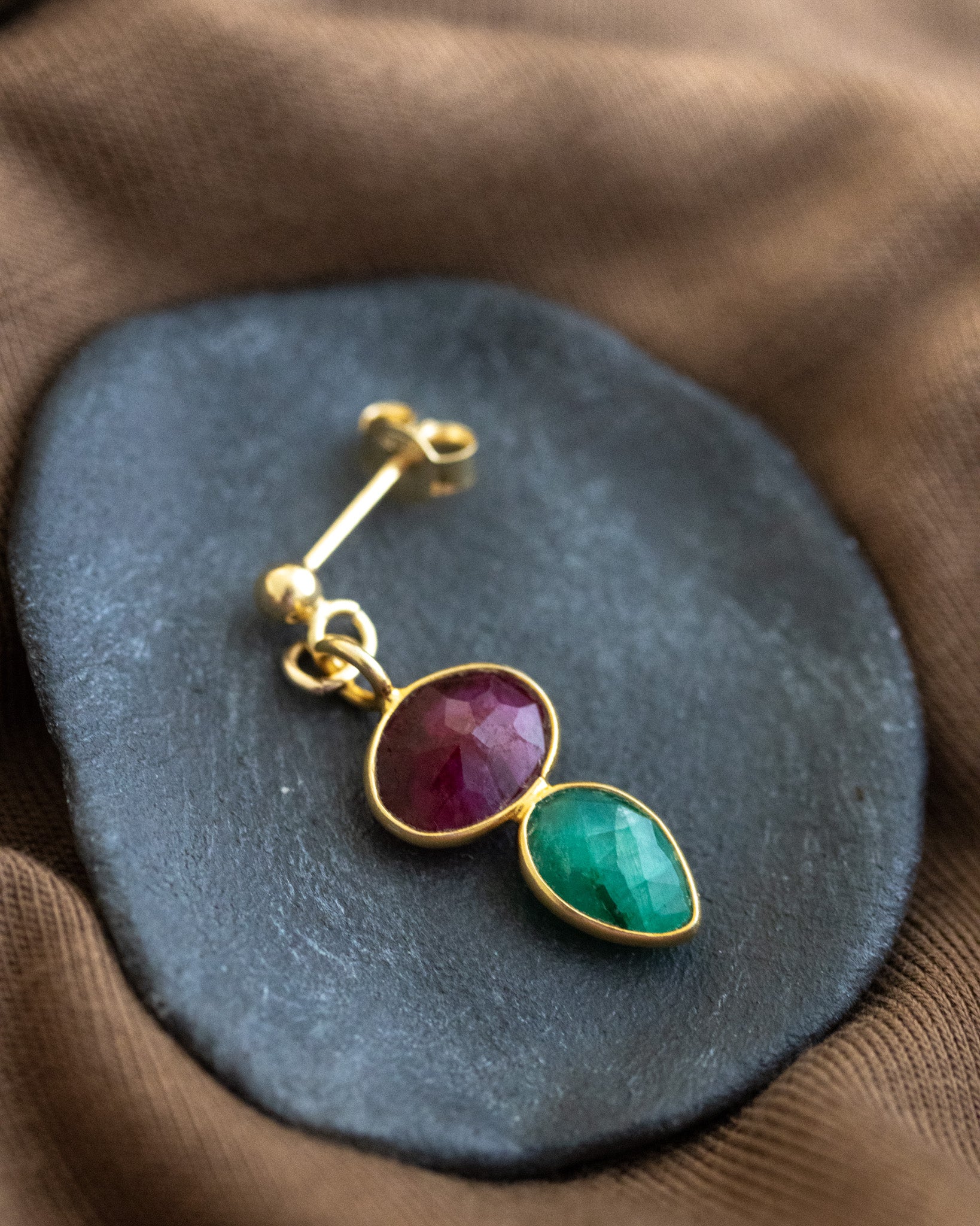 Ein Ohrstecker, der aus einem gold gerahmten Rubin und einem darunter angebrachten, ebenfalls gold gefassten, Smaragd besteht, liegt auf einem dunkelgrauen Steinschälchen.