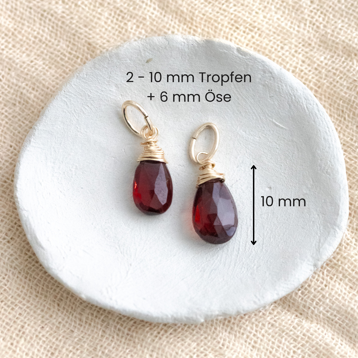 Zwei gold gefasste Granat-Anhänger mit Beschriftung zu Maßen auf einem Schälchen ausgelegt.