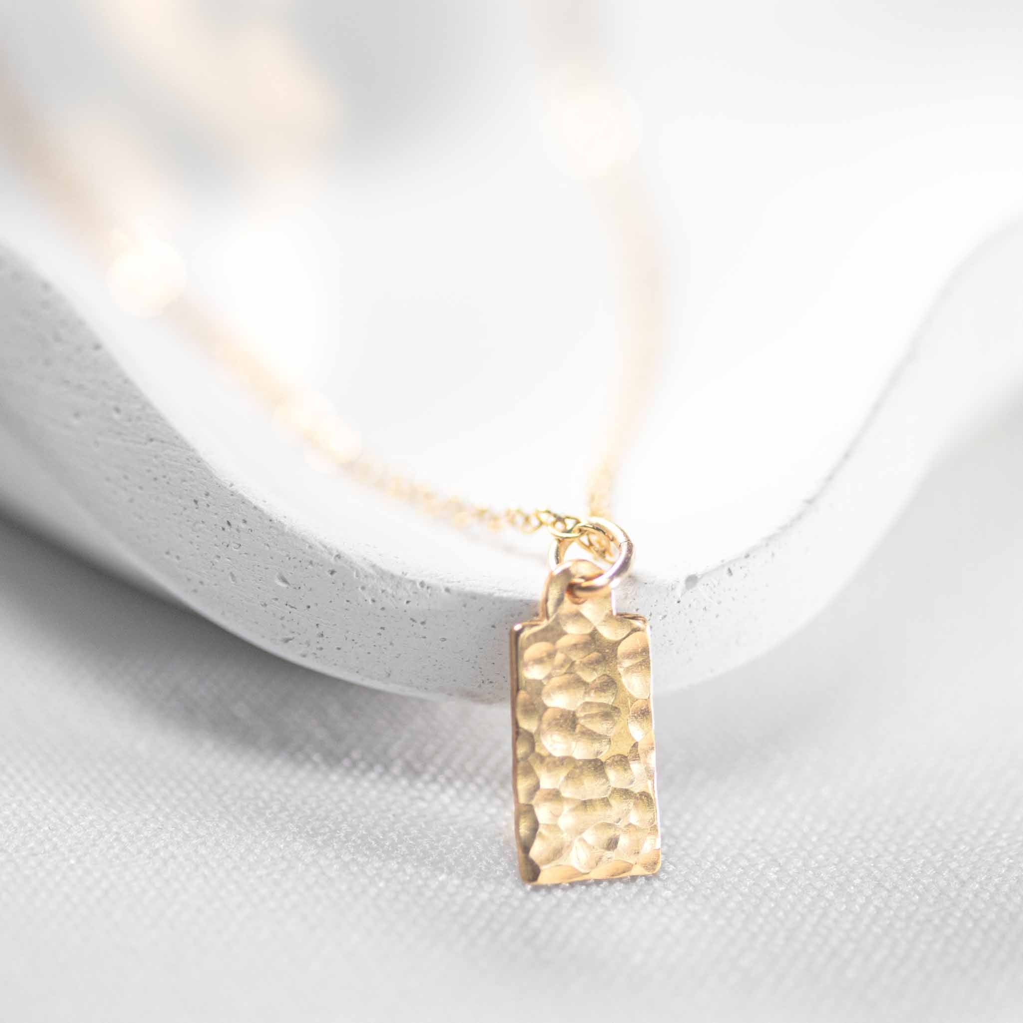Eine feine und kurze Goldkette mit einem rechteckigen, gehämmerten Plättchen-Anhänger lehnt an einem weißen Schälchen.