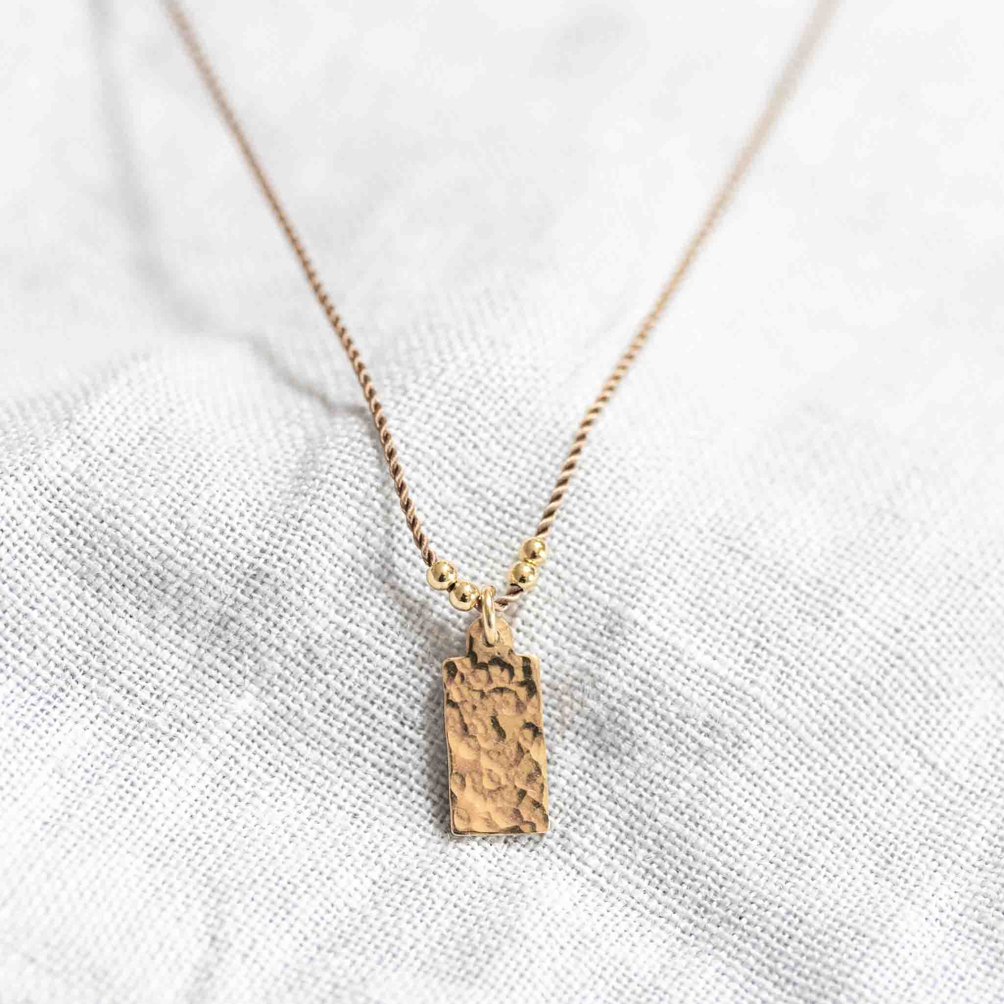 Eine feine, beige Kordelkette mit einem rechteckigen, gehämmerten Plättchenanhänger auf weißem Hintergrund.