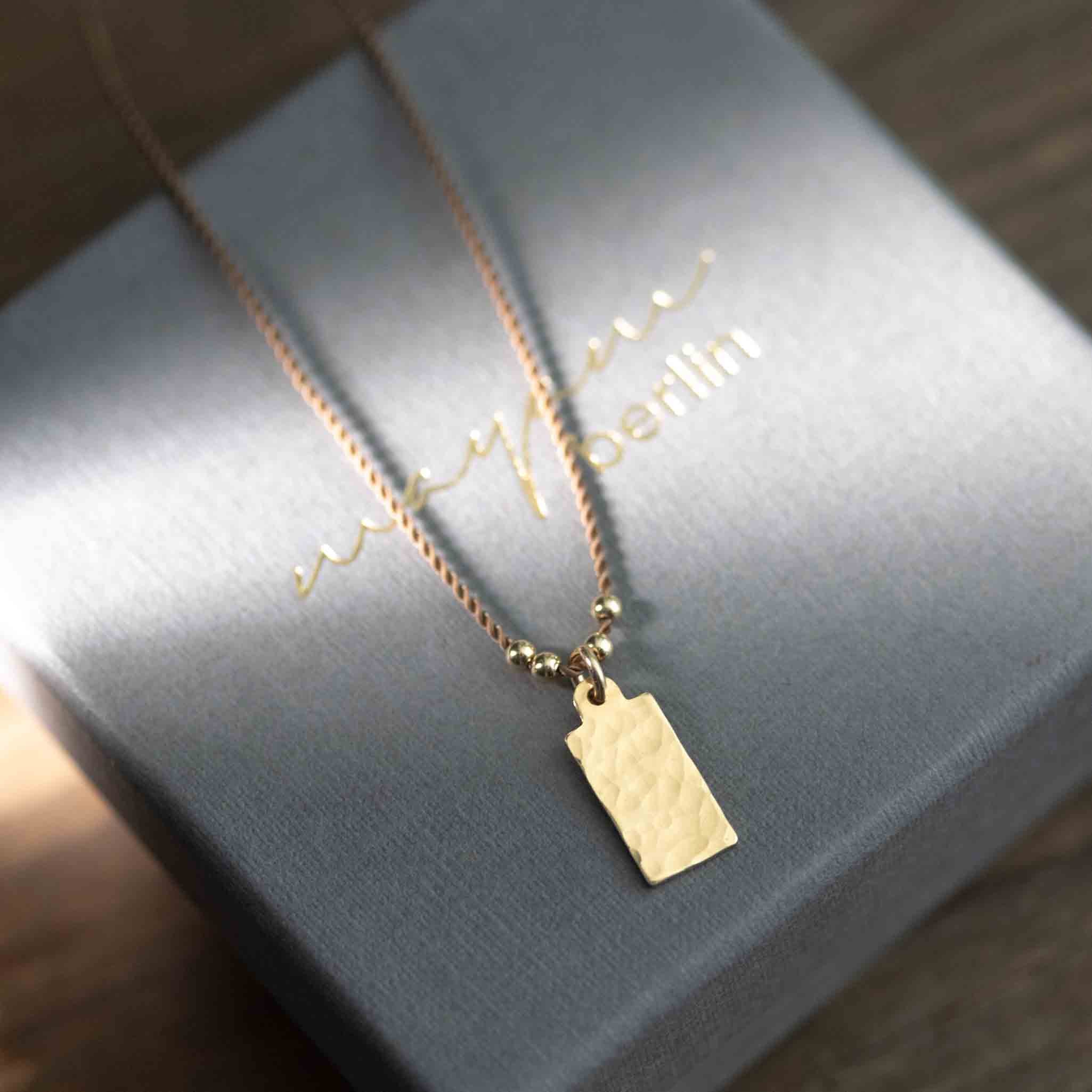 Eine feine, beige Kordelkette mit einem rechteckigen, gehämmerten Plättchenanhänger auf einem grauen Karton-Etui.