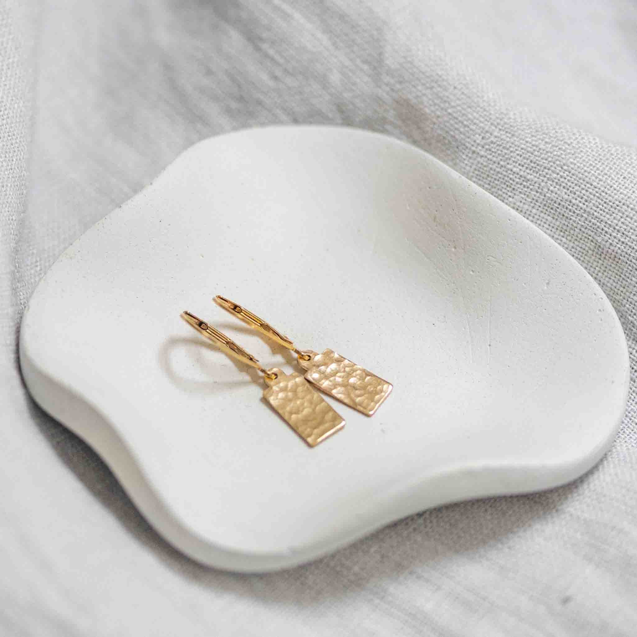 Ein Paar goldener Ohrringe, bestehend aus Klappbrisuren und gehämmerten Plättchen liegt in einem weißen Schälchen.