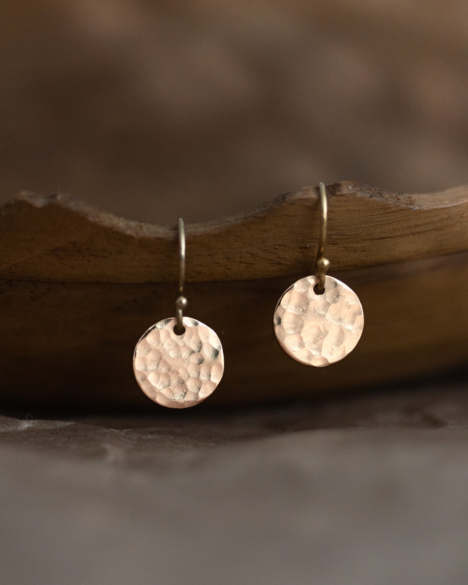 Ein Paar goldener Plättchenohrringe hängt an einer gewellten, dunkle Holzschale. Die runden Plättchen sind gehämmert.