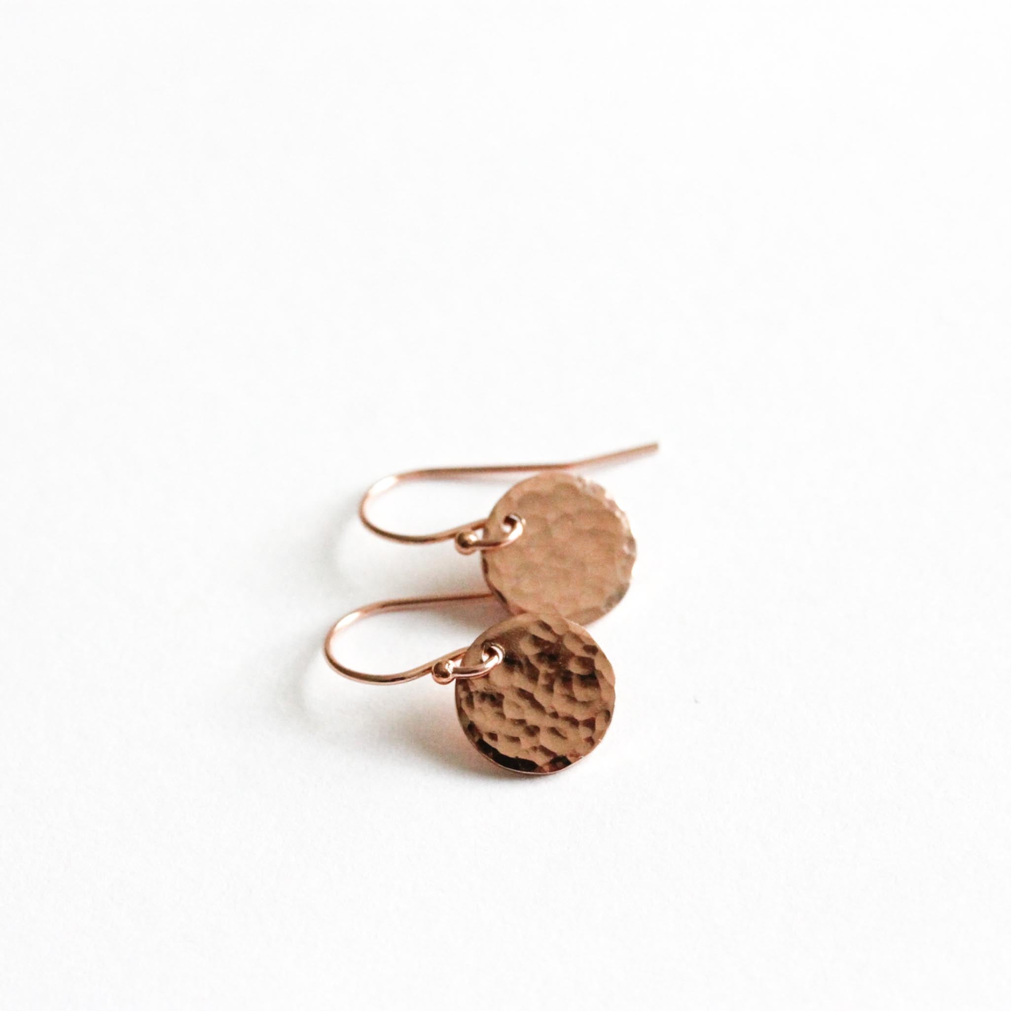 Auf weißem Untergrund liegt ein Paar rosé goldener Ohrringe. An den Ohrhaken hängt jeweils ein rundes, gehämmertes 9 mm Plättchen.