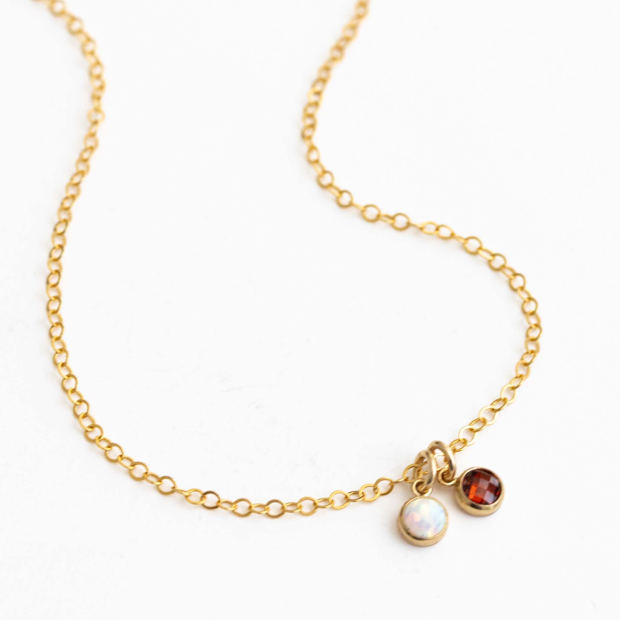 Eine zierliche Goldkette mit zwei winzigen, runden Anhängern, einem Opal und einem Granat, auf weißem Hintergrund.
