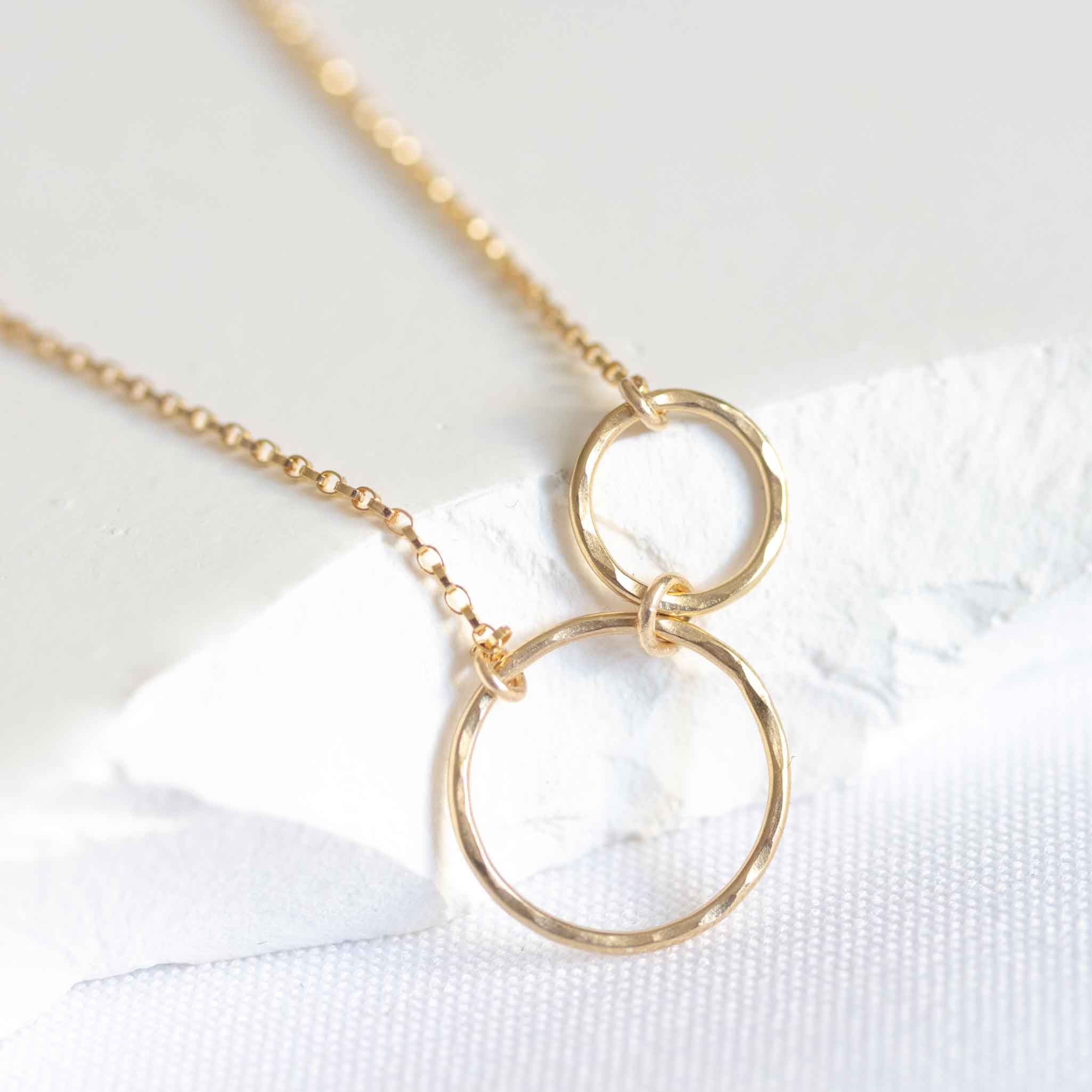 Zwei unterschiedlich große Ringe mit gehämmerter Struktur sind in ein dünnes Goldarmband eingearbeitet und lehnen an einem weißen Stein.