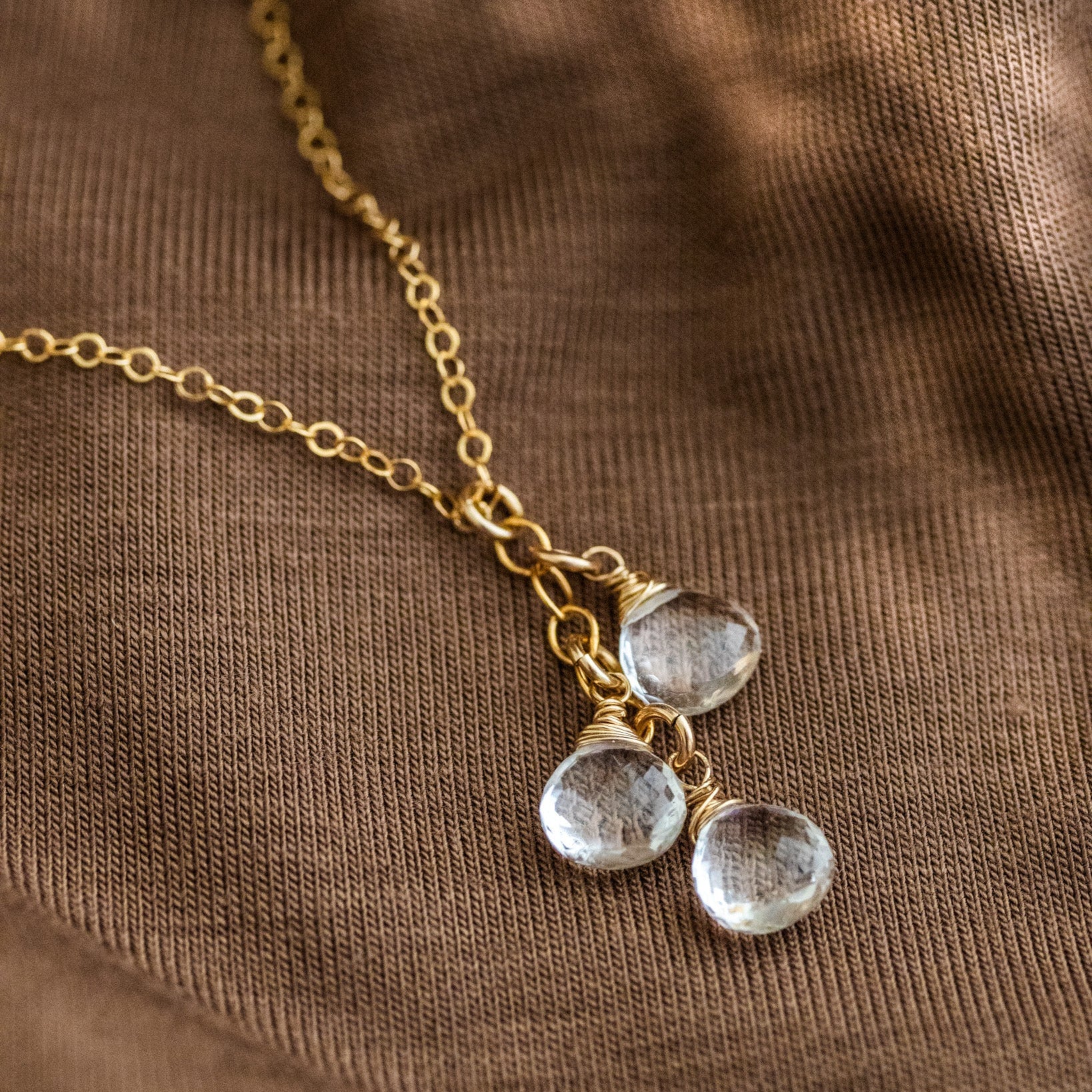 Eine feine Goldkette mit drei untereinander angebrachten, kleinen Bergkristalltropfen liegt auf braunem Stoff.