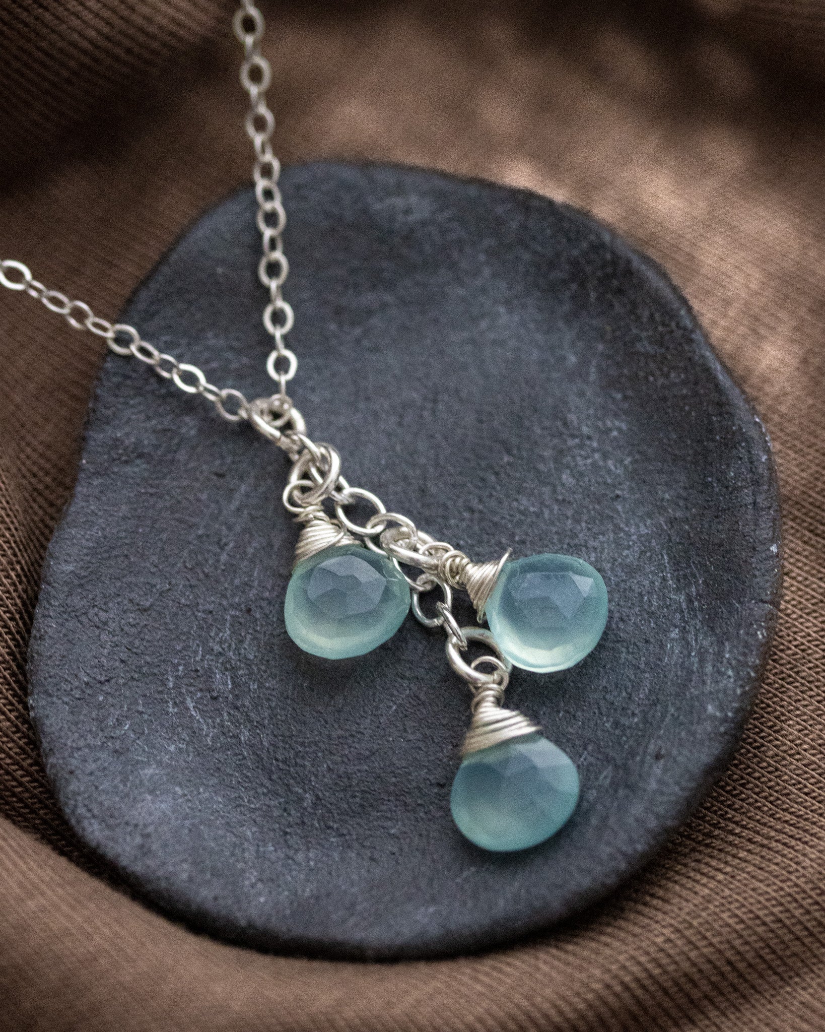 Eine dünne Silberkette mit drei untereinander angebrachten Aqua Chalzedon-Tropen liegt auf einem grauen Steinschälchen.