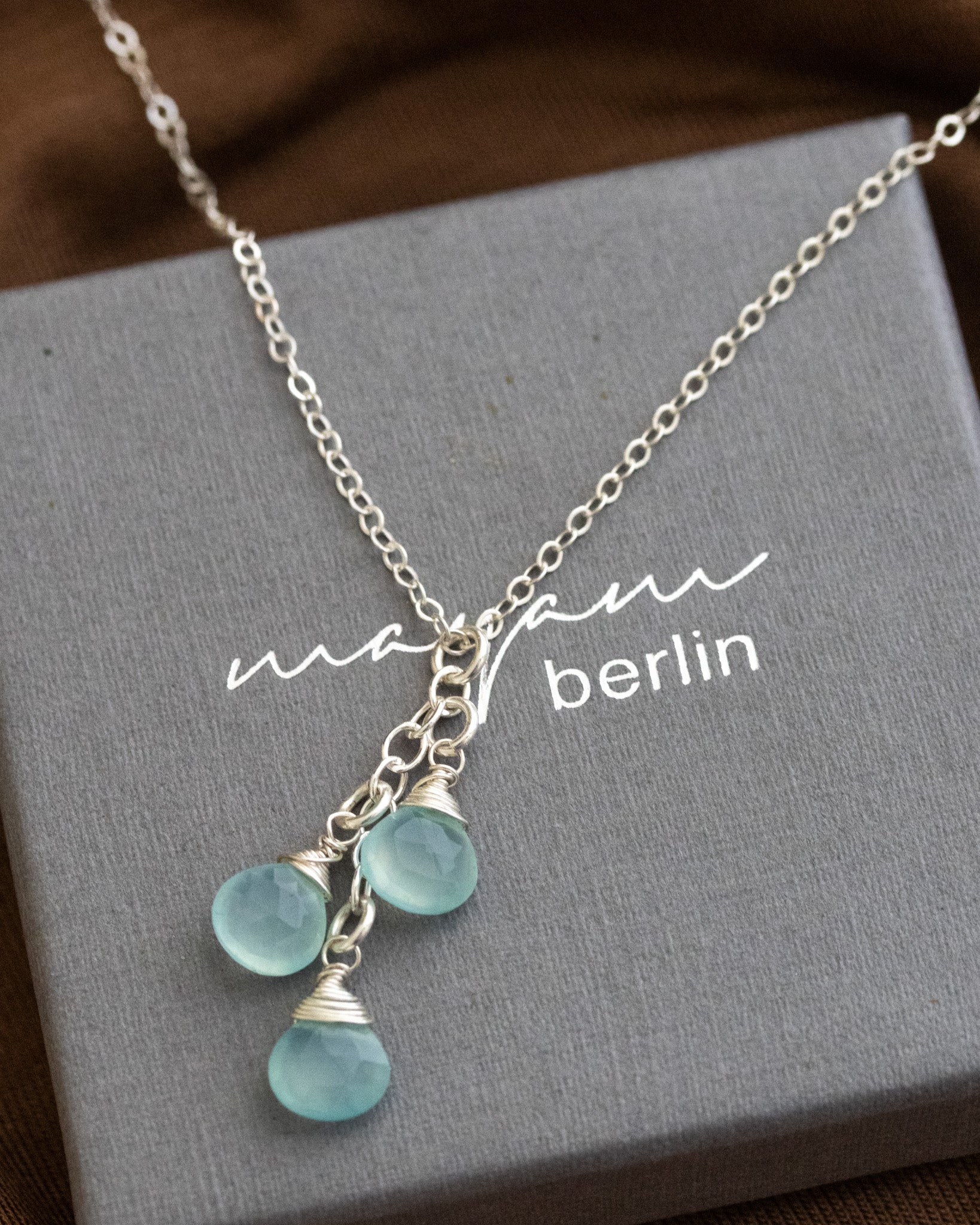 Eine dünne Silberkette mit drei untereinander angebrachten Aqua Chalzedon-Tropen liegt auf einem grauen Schmucketui.