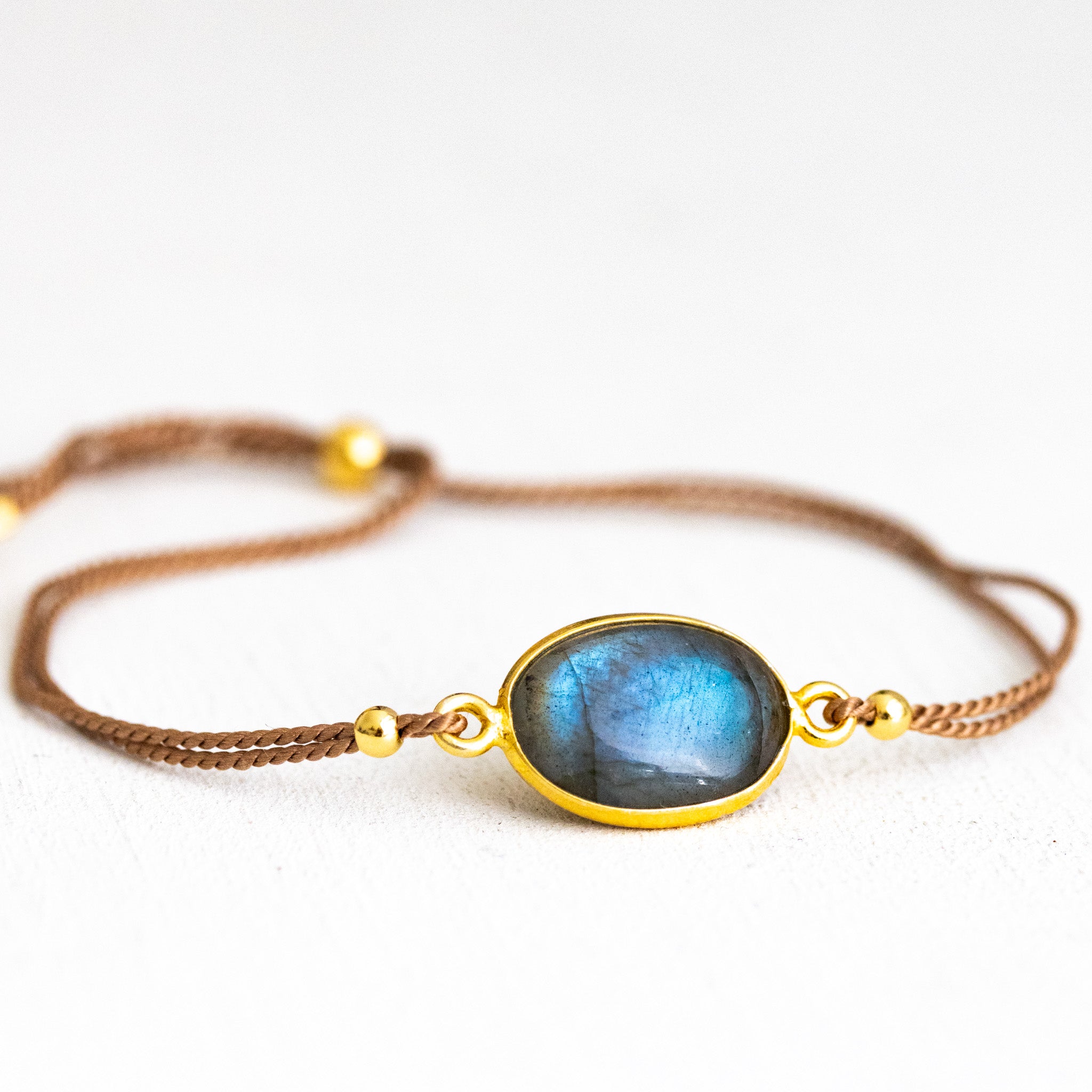 Ein dünnes, beiges Seidenarmband mit einem ovalen und in gold gefassten Labradorit in der Mitte liegt auf weißem Hintergrund.