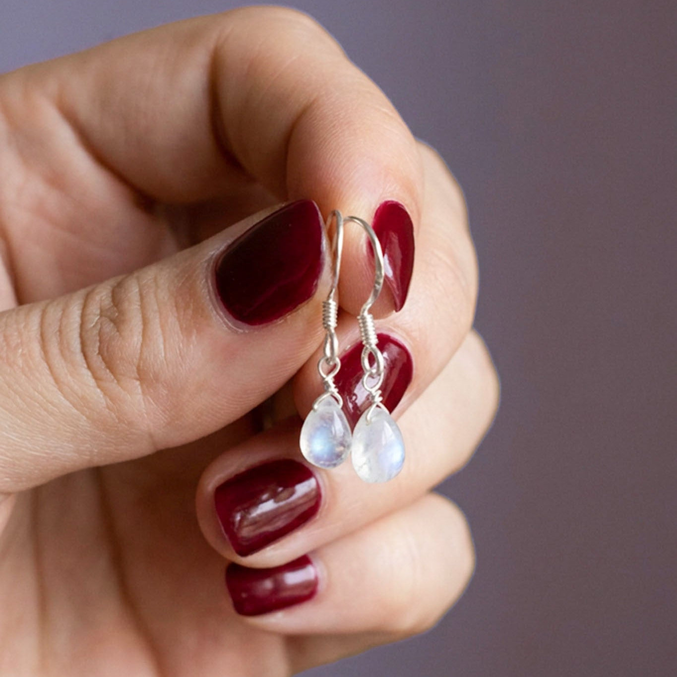 Eine Hand mit rot lackierten Fingernägeln hält ein Paar Silber-Ohrringe mit Mondstein-Tropfen.