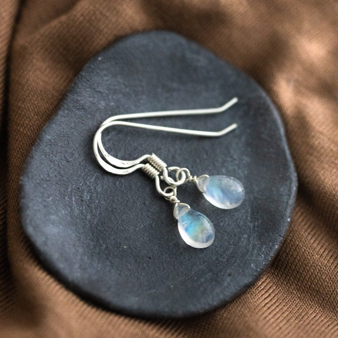 Ein Paar Silber-Ohrringe mit kleinen Mondstein-Tropfen liegt auf einem schwarzen Schälchen auf braunem Stoff.