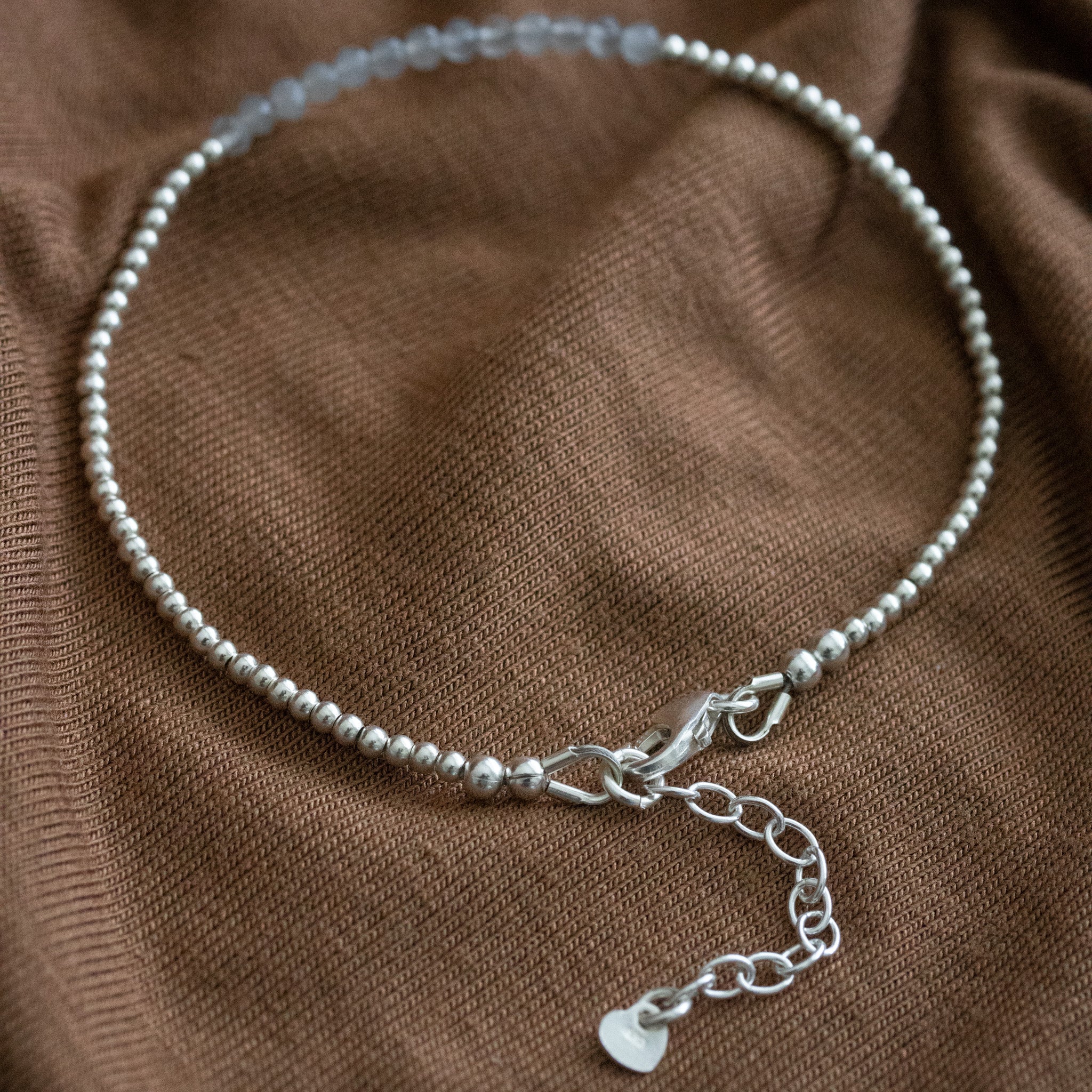 Ein ultra dünnes Silberarmband mit grauen Mondsteinperlen in der Mitte liegt auf braunem Stoff. 